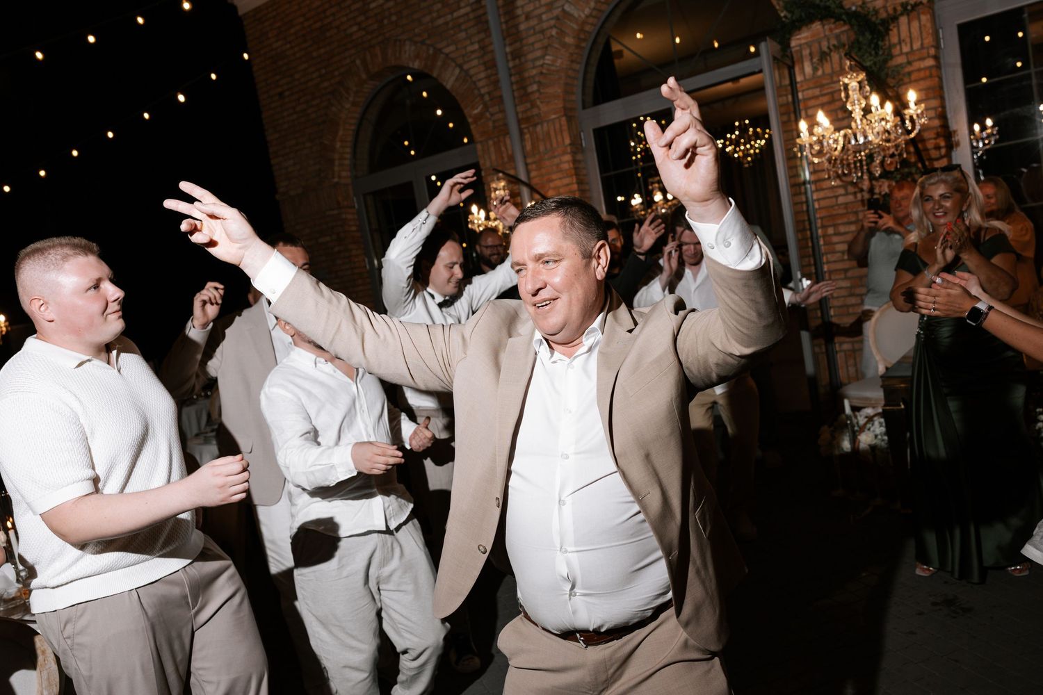 Wedding reception guests dancing and celebrating under dimly lit chandeliers.