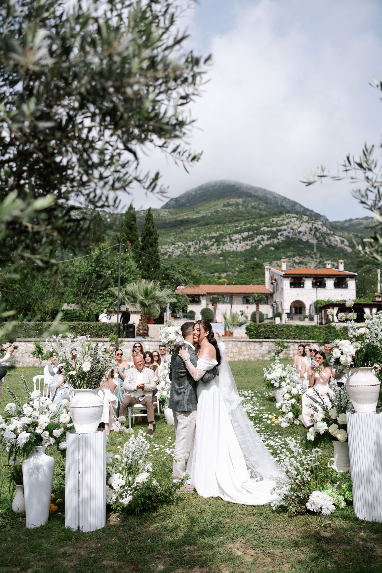 Romantic outdoor wedding ceremony in an Italian garden with white floral arrangements and mountain backdrop.