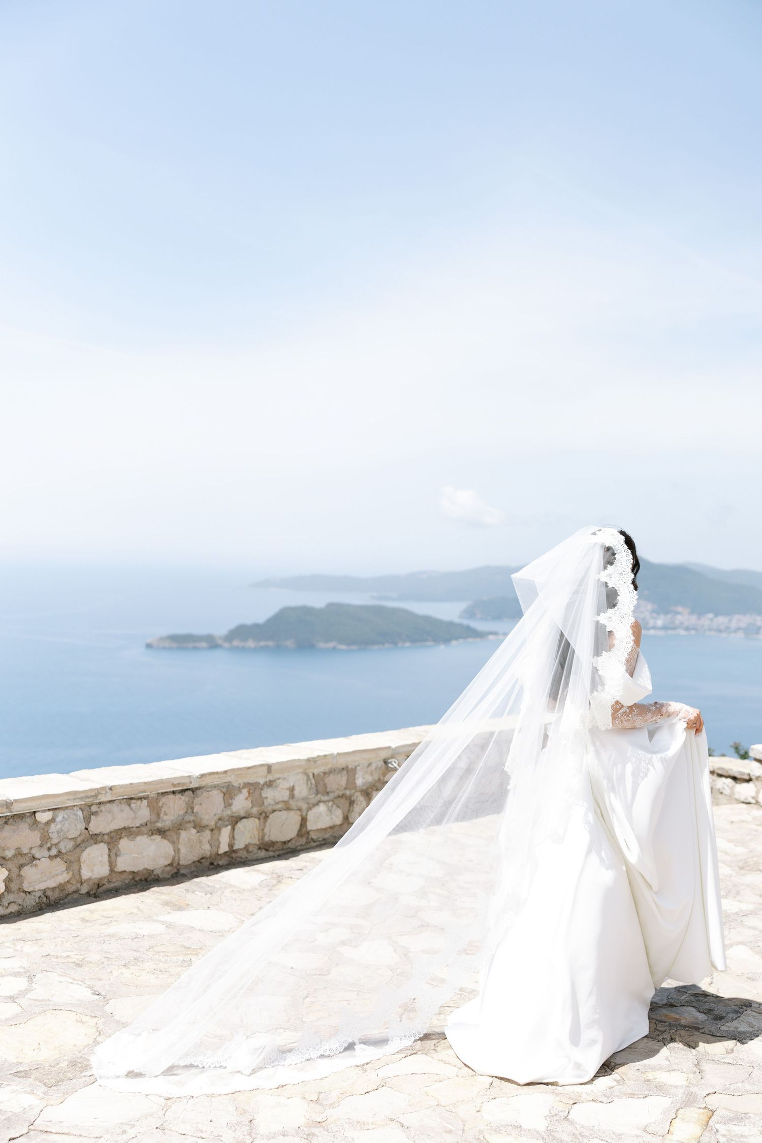 A bride in a flowing white gown and long veil stands on a stone terrace overlooking a coastal Mediterranean landscape.