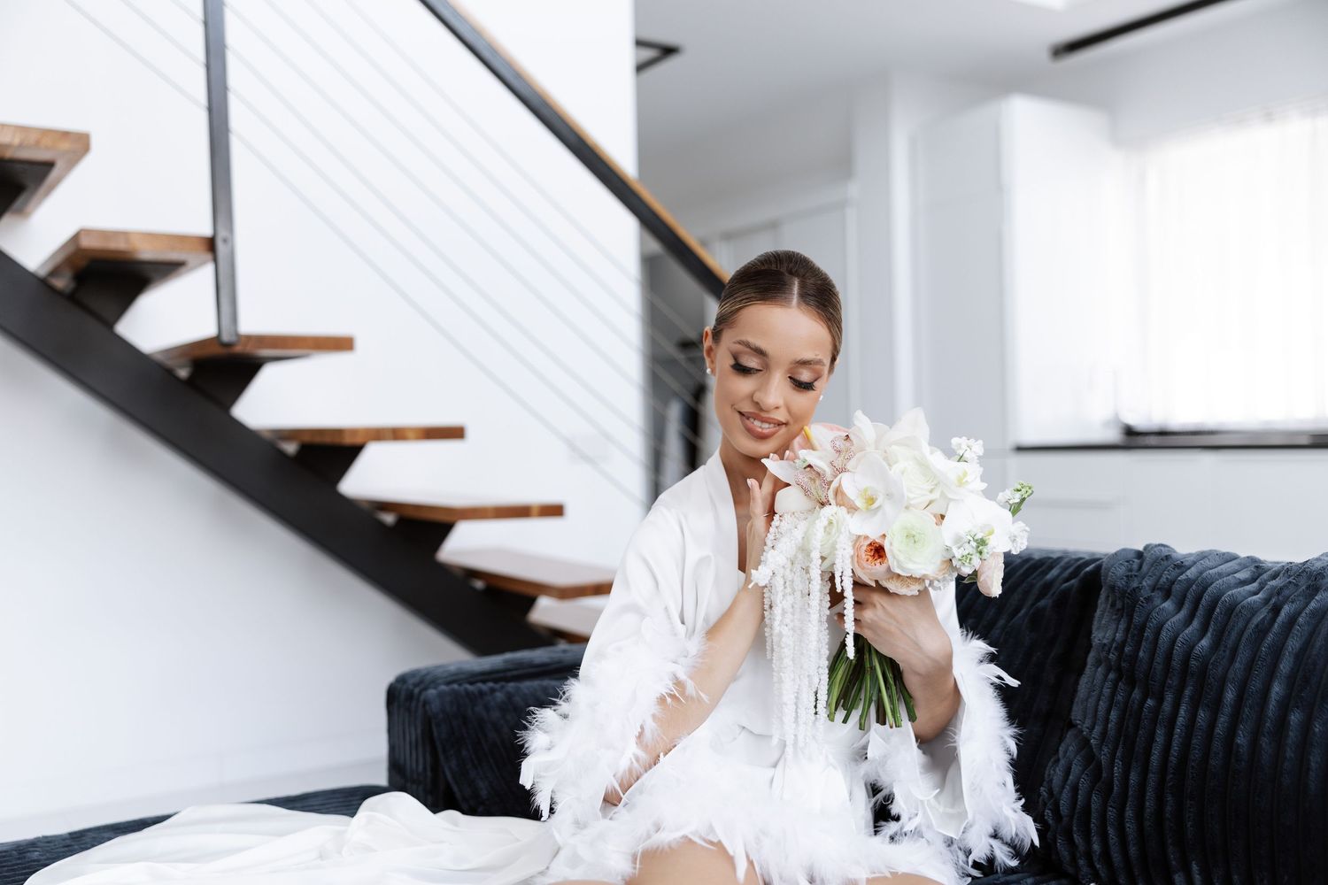 A dancer in white reclines on a navy velvet couch while holding white orchids beneath a modern staircase.