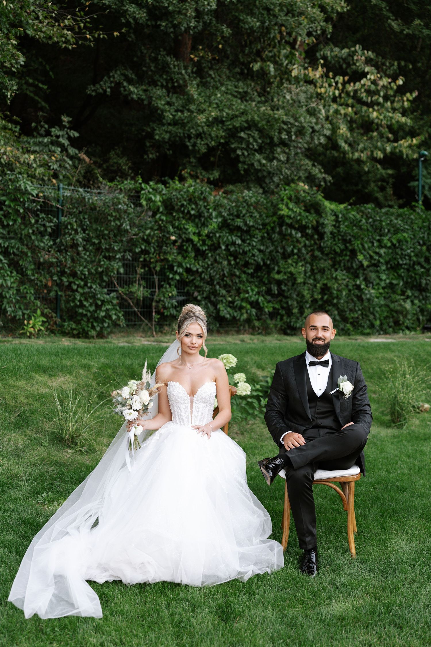 A couple in wedding attire sits on wooden chairs on a lush green lawn during an outdoor photo session.
