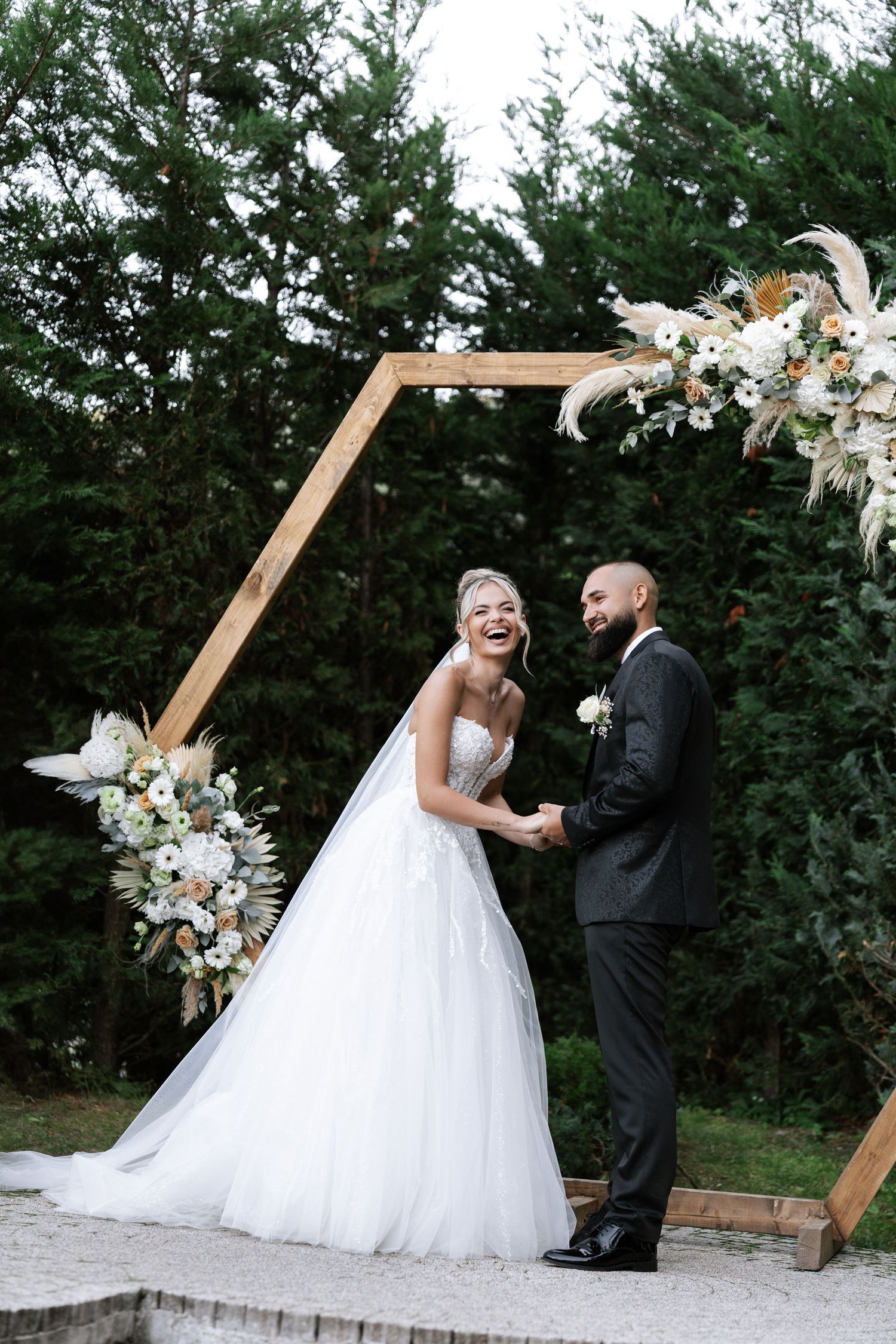 A couple shares a joyful moment under a wooden hexagonal arch decorated with white and peach floral arrangements.