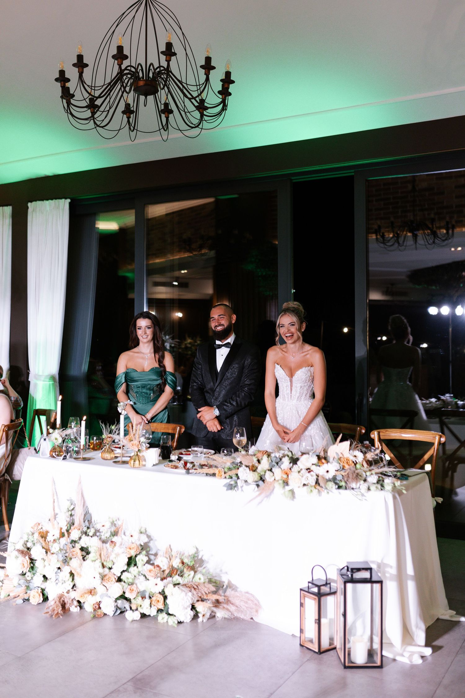 Elegant wedding reception head table decorated with lush white floral arrangements and black chandeliers overhead.