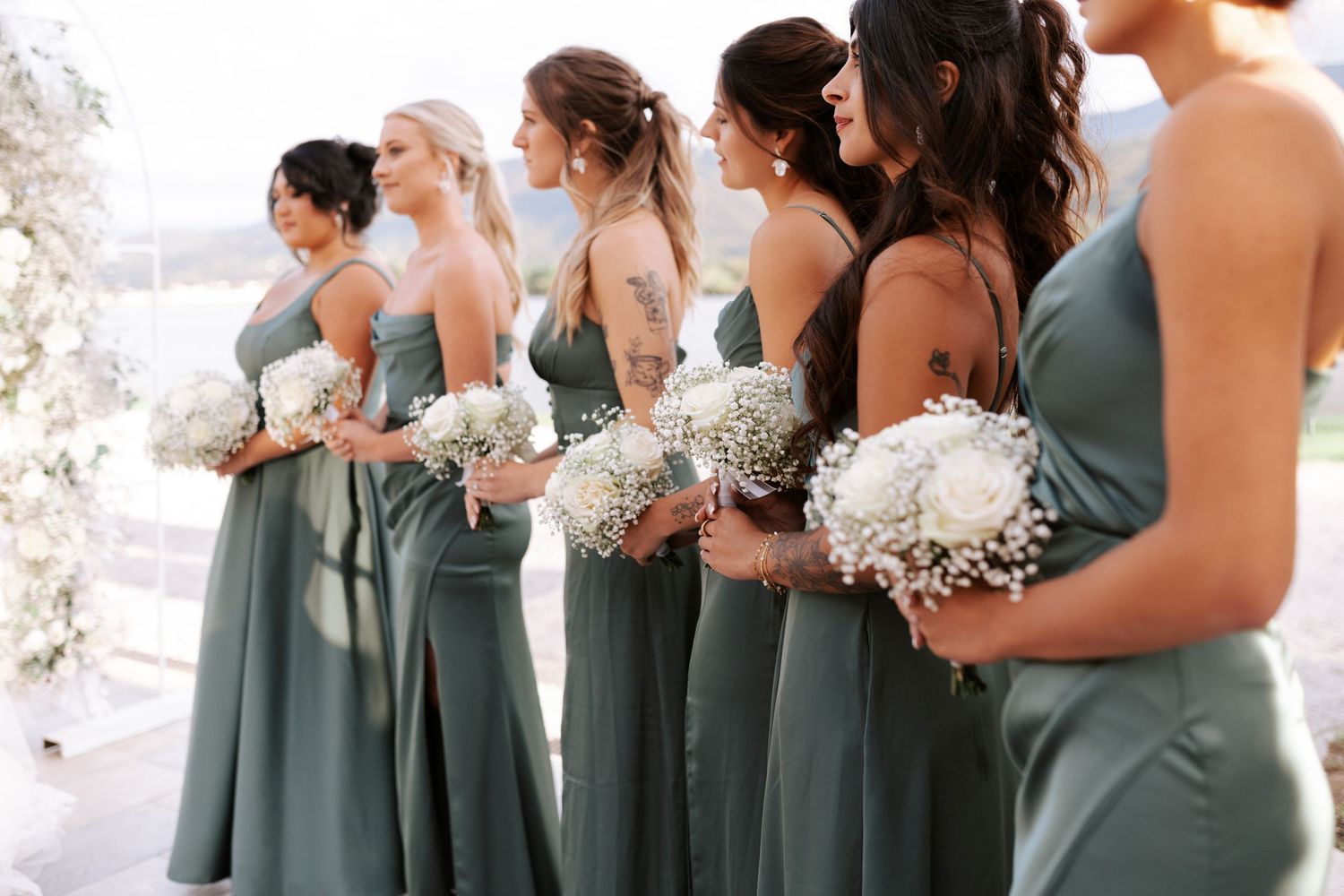 Bridesmaids in sage green dresses hold white bouquets at outdoor ceremony.