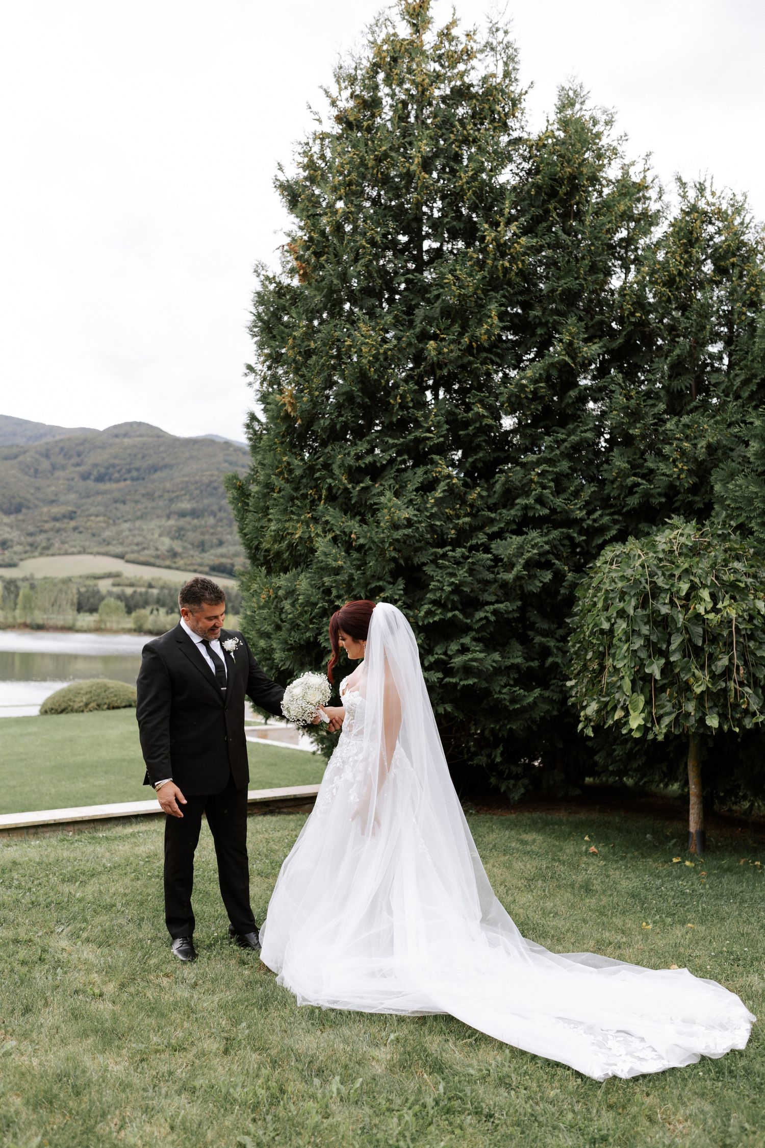 Wedding couple poses in front of evergreen trees with lake and mountains in background.