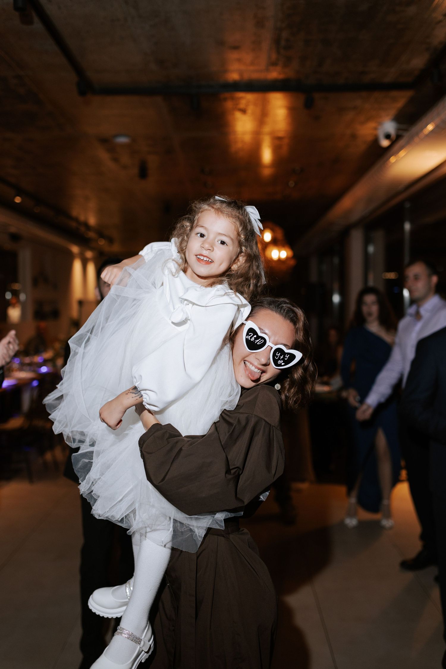 A flower girl in white dress being carried at a wedding reception while wearing heart sunglasses.