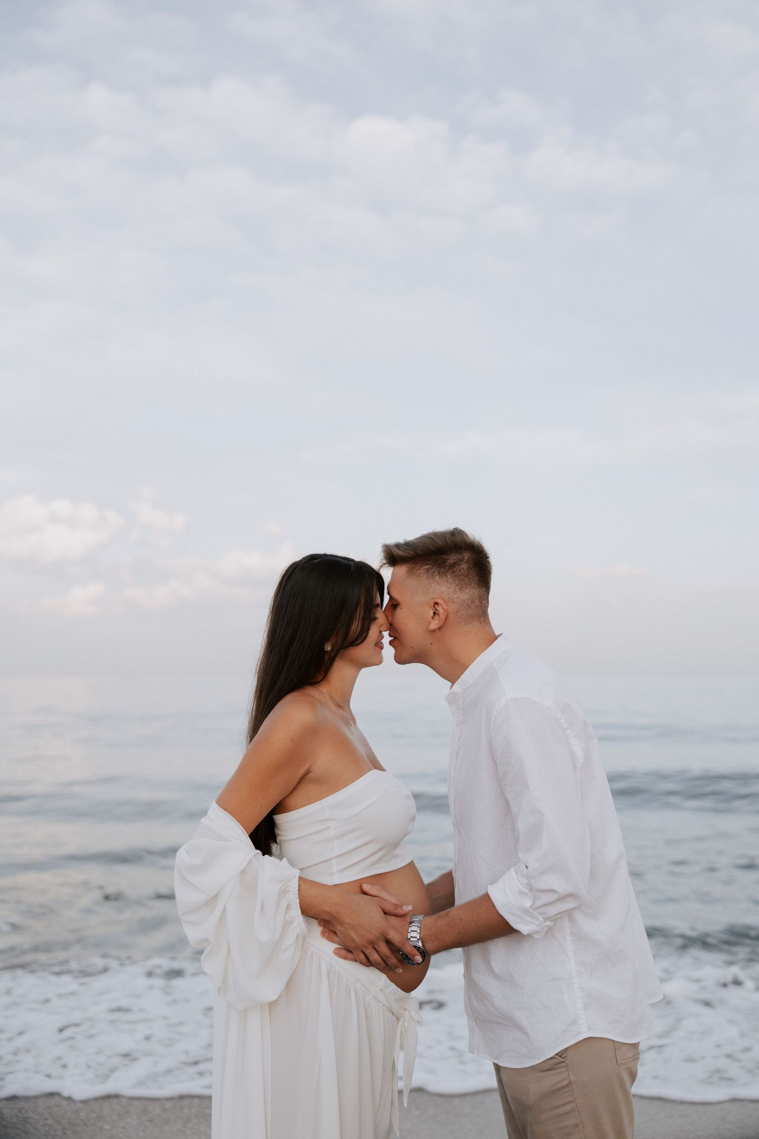A romantic maternity photoshoot on the beach at sunset with a couple in white and beige clothing.