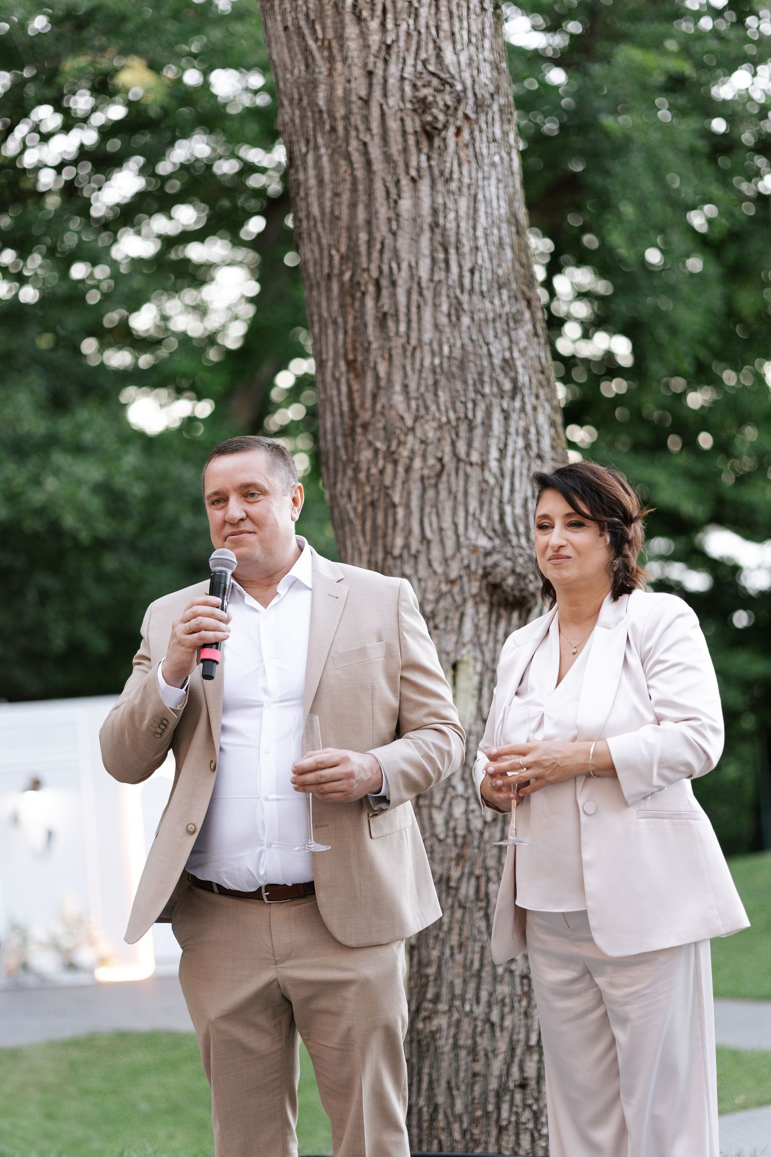 Wedding guests in beige suits making toasts outdoors among tall trees.