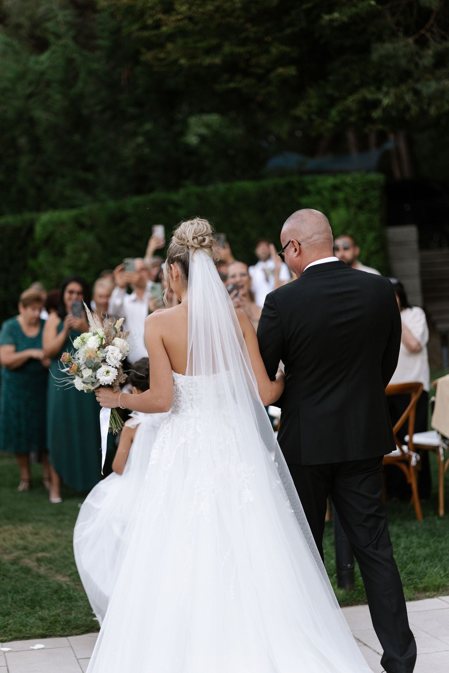 A bride in a flowing white wedding dress and groom in black tuxedo dance together at an outdoor wedding reception.