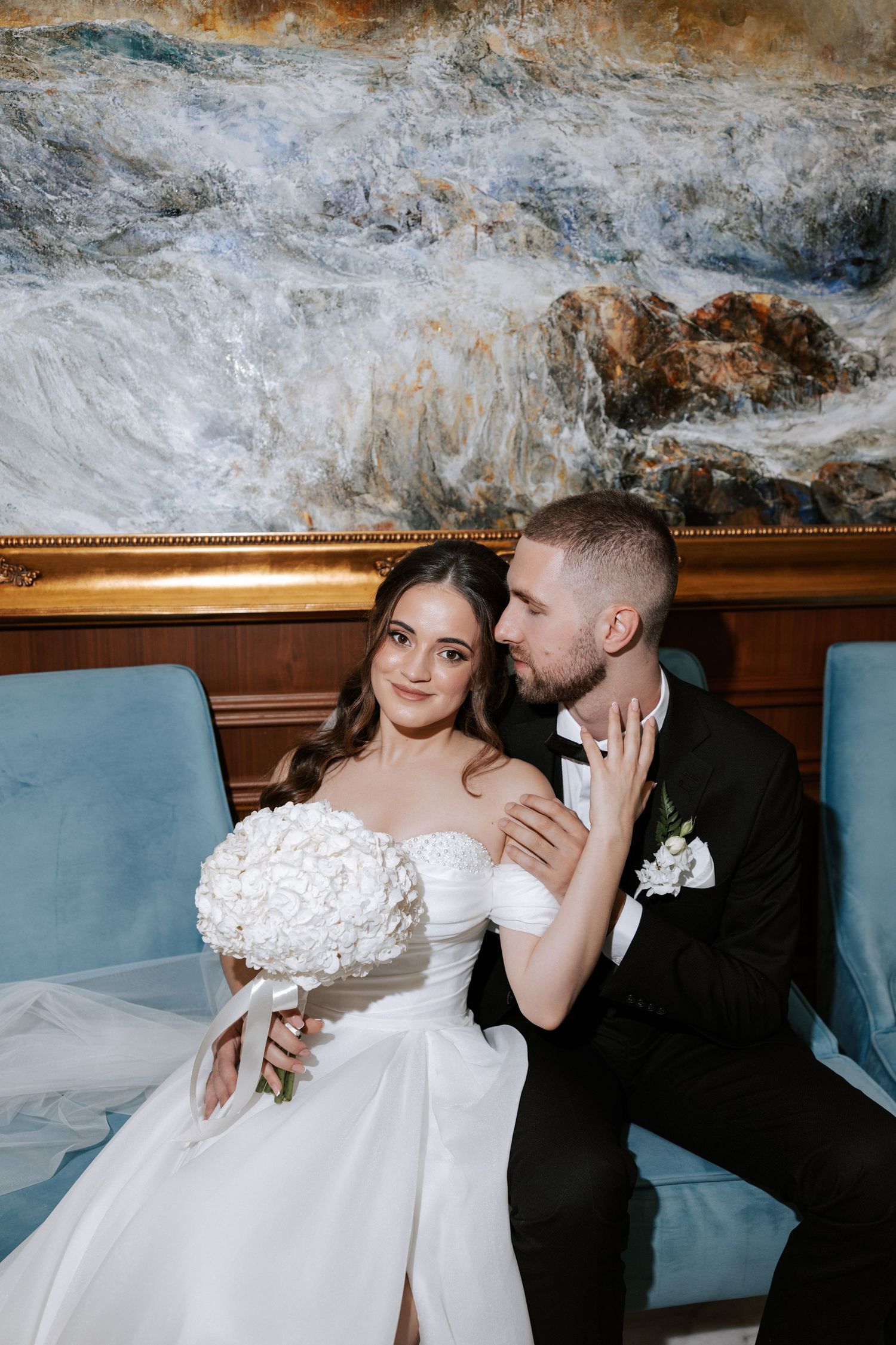 Wedding couple poses on blue chairs against dramatic seascape artwork in ornate gold frame.