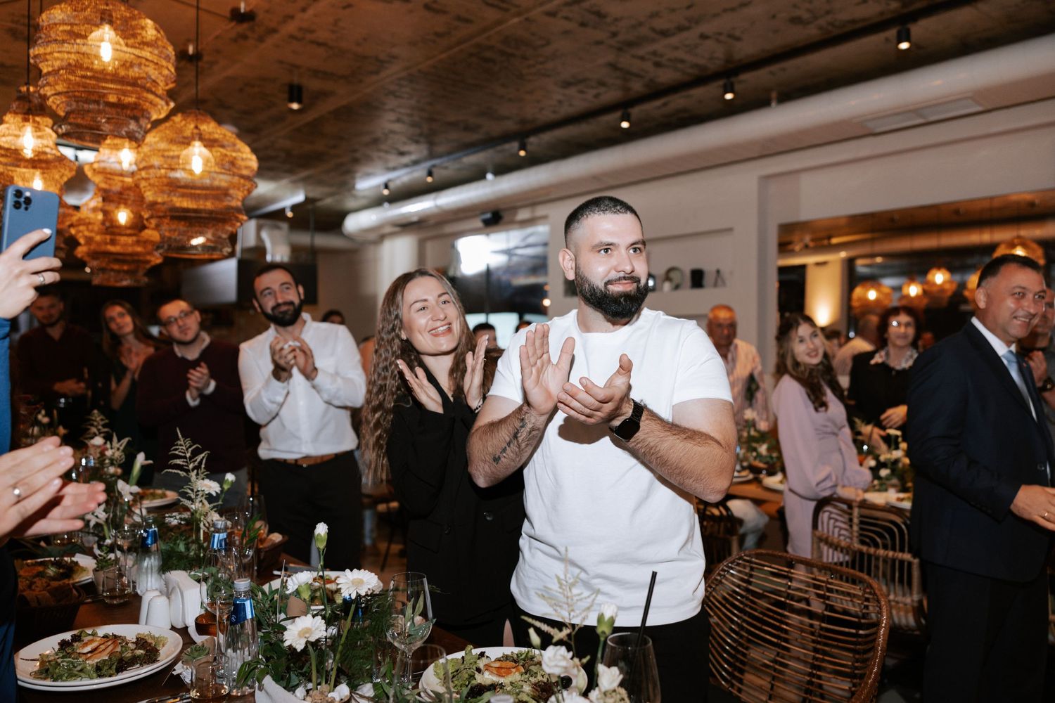 Wedding guests applauding during a reception celebration in a modern restaurant setting with ambient lighting.