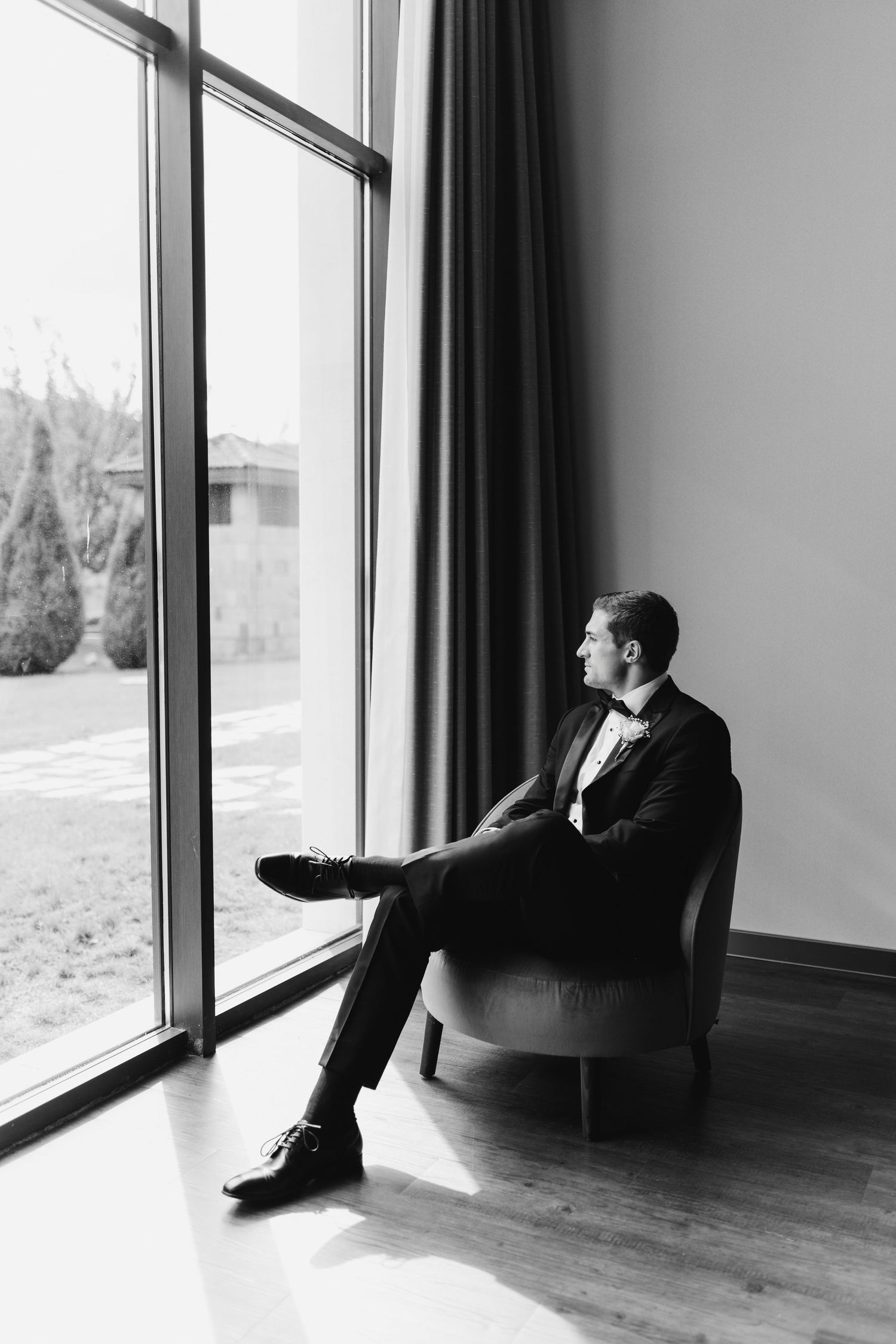 A black and white photo of someone in a tuxedo sitting in an armchair by a large window.