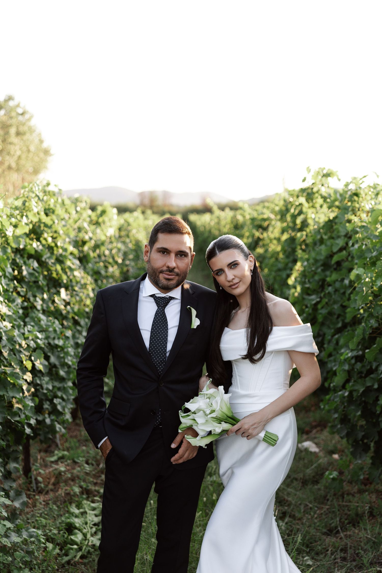 A couple poses together in a vineyard wearing formal wedding attire against a backdrop of green grapevines.