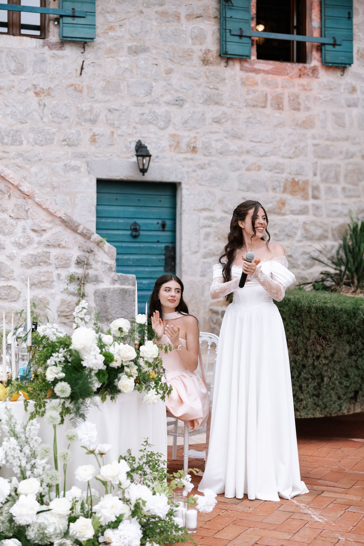 Wedding party poses on rustic terrace with stone walls, teal shutters and elegant white floral arrangements.