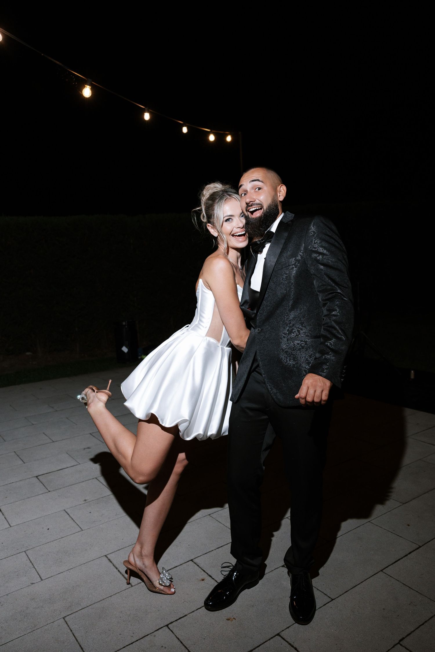 A couple in formal attire dances together at night under string lights on a stone patio.