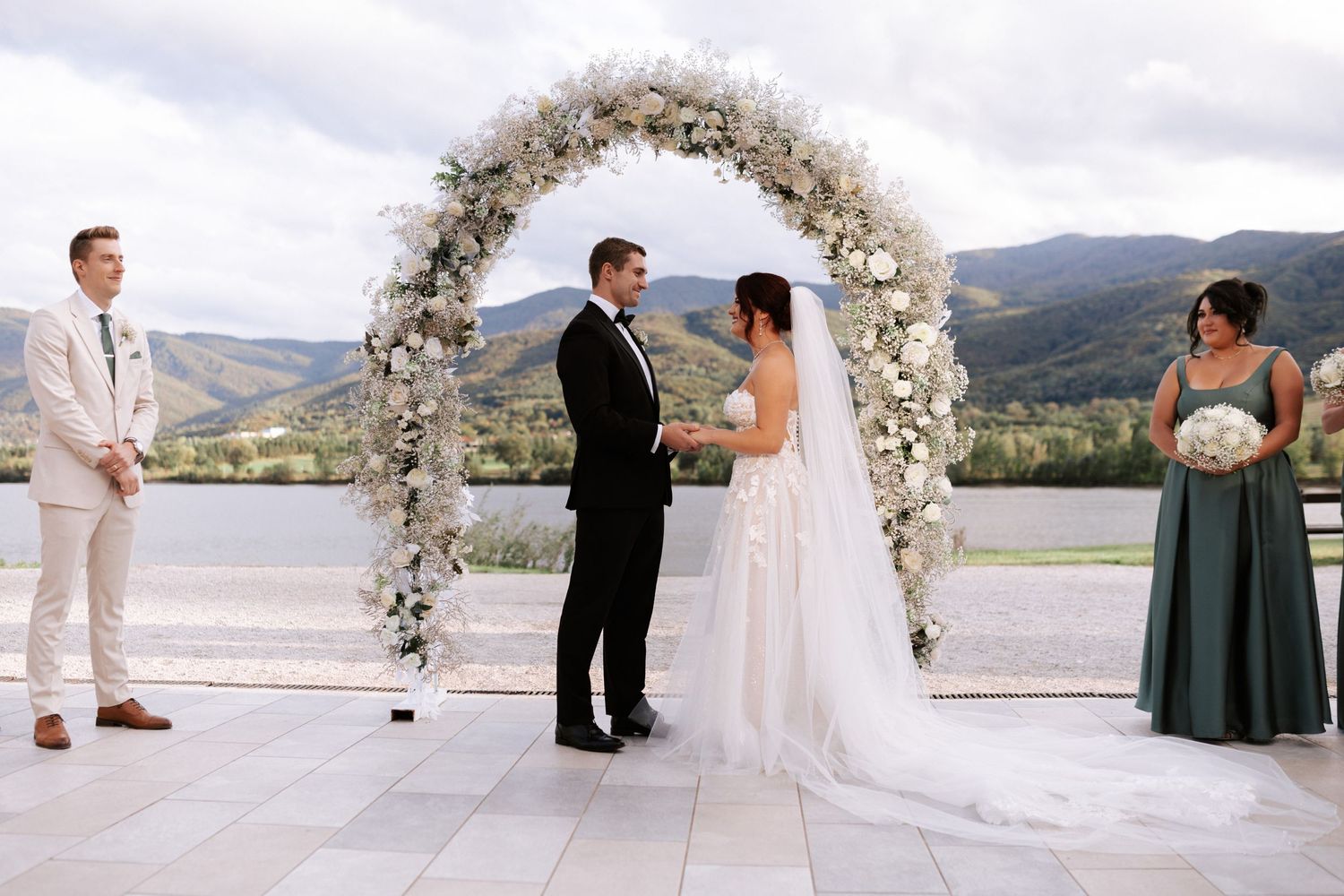 Wedding ceremony under a white floral arch with mountain views and guests looking on.