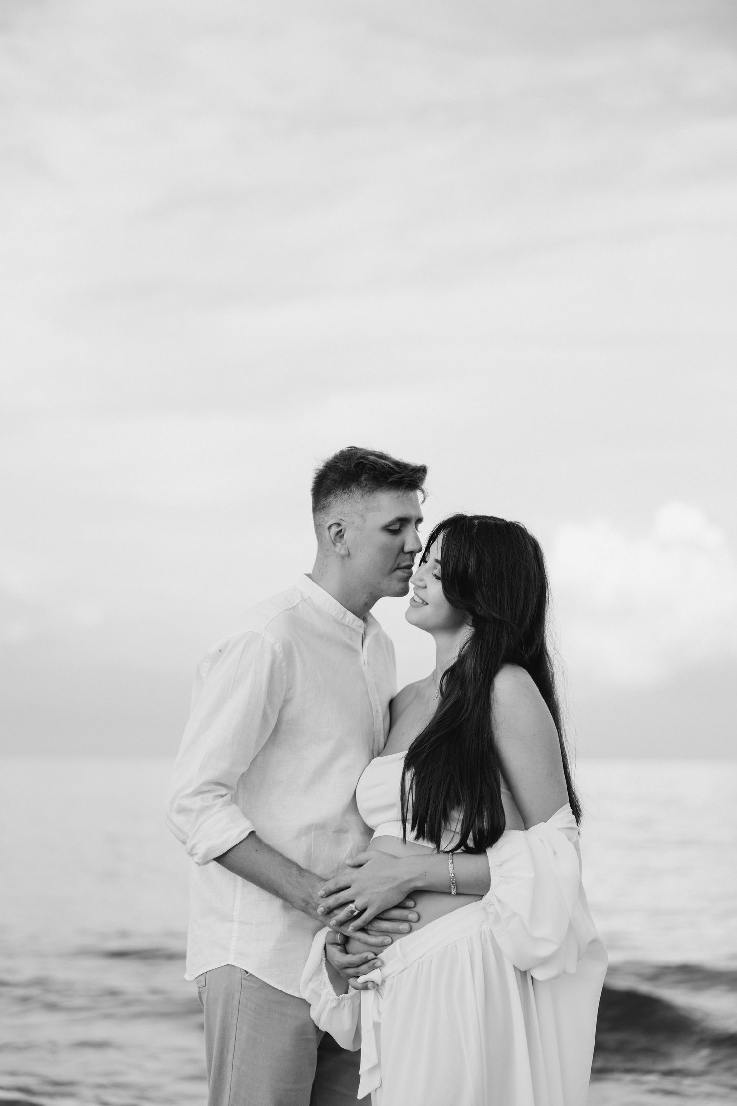 A black and white romantic beach photo of an expecting couple in white attire.