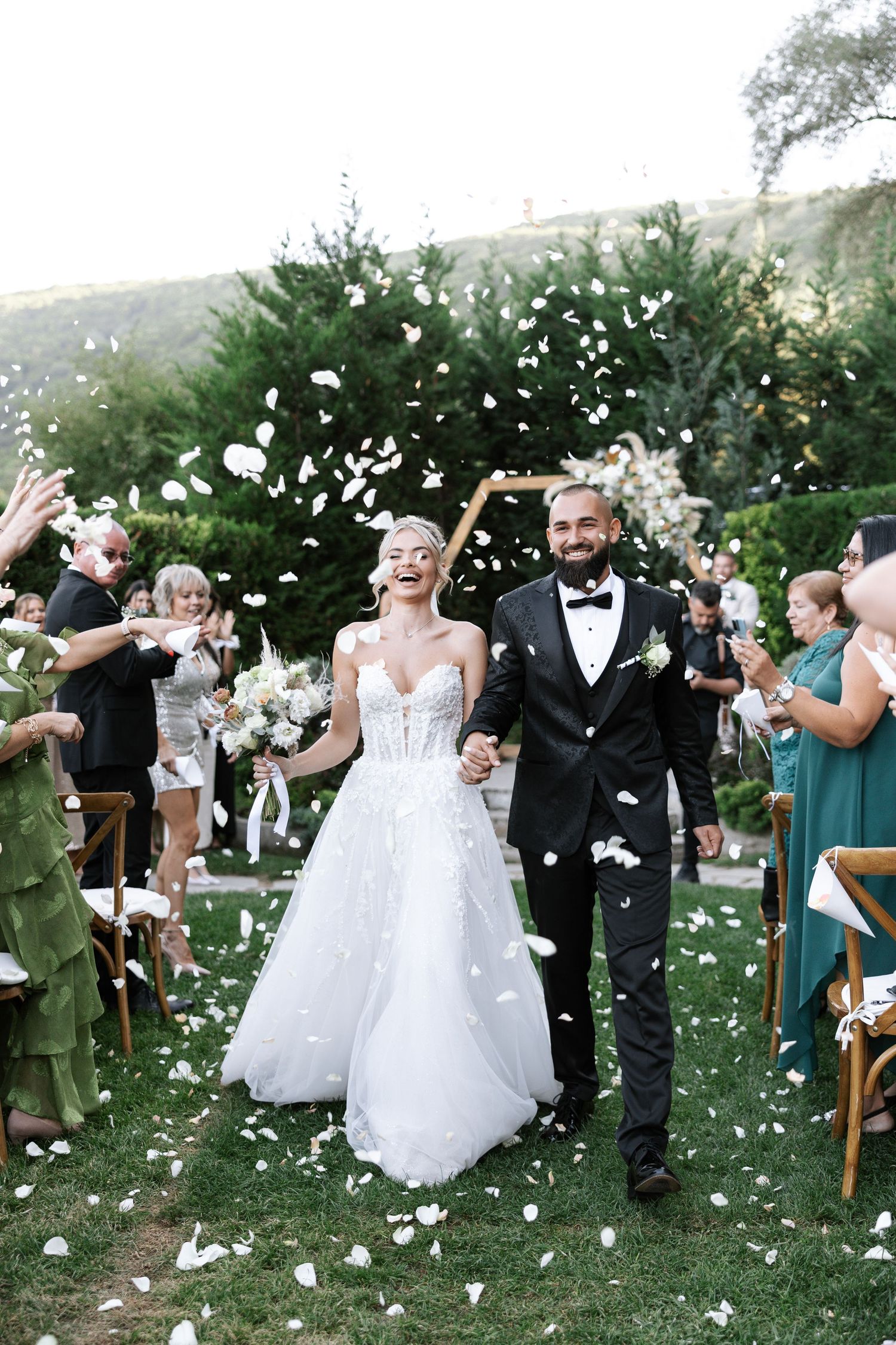 A wedding couple walks down an outdoor aisle while guests throw white flower petals in celebration.