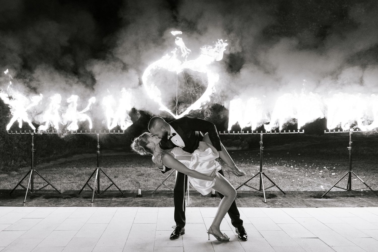 A couple performs a romantic dance dip pose against a dramatic backdrop of fire and smoke at an outdoor event.