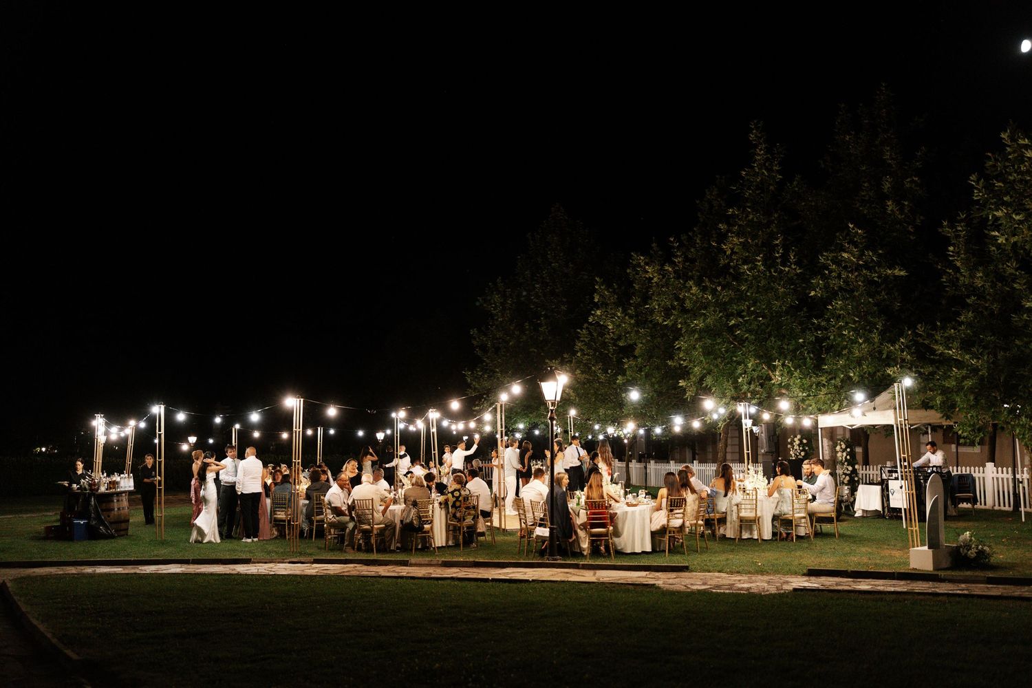 String lights illuminate an outdoor evening event with tents and people gathering in a park setting.