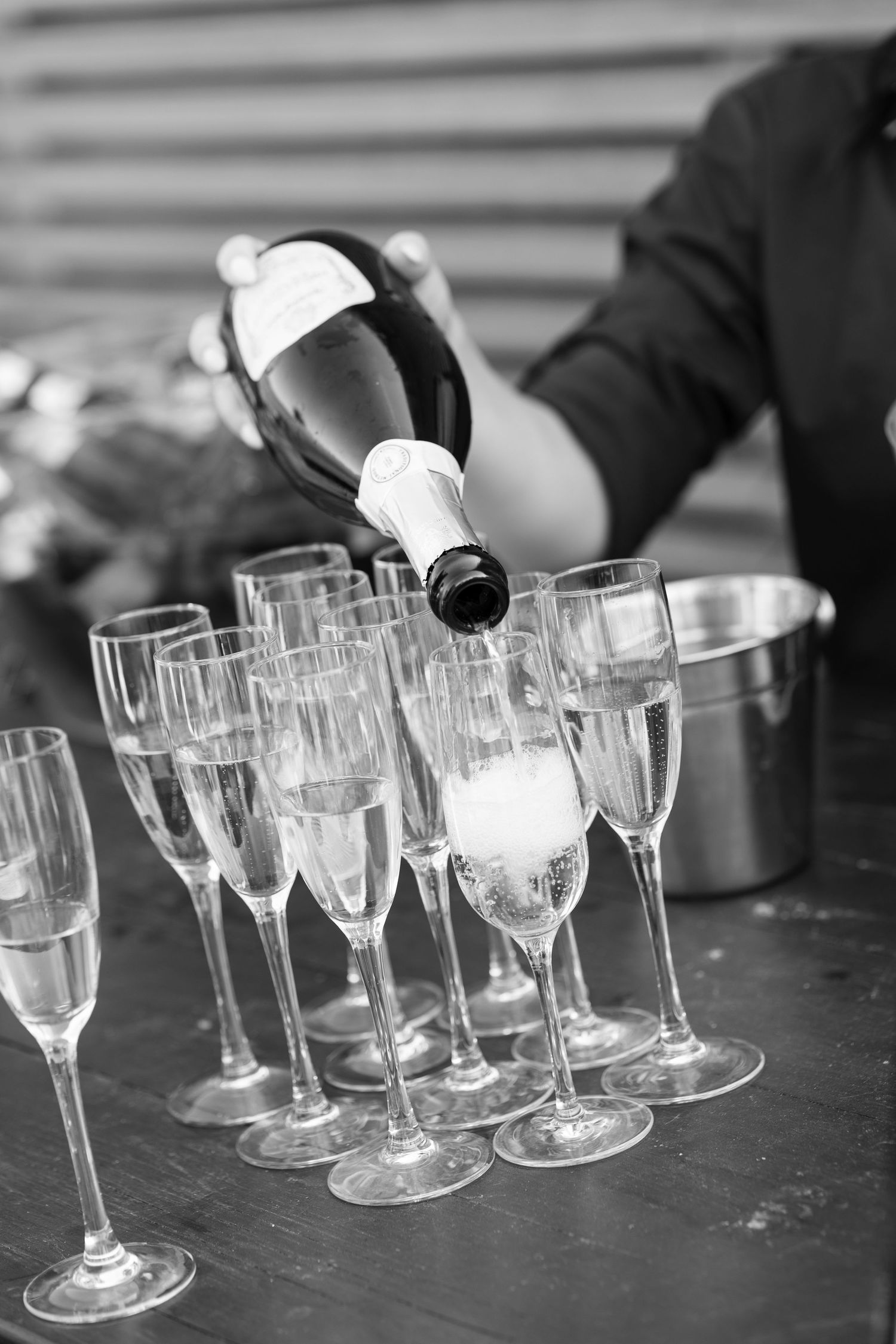 A person pours champagne into elegant flute glasses arranged on a dark surface with an ice bucket in the background.