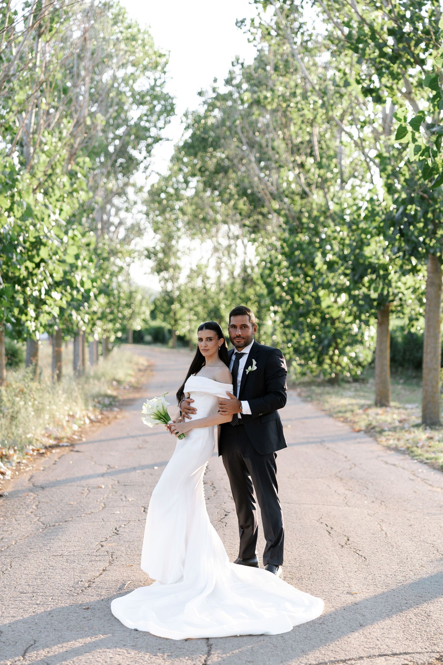 A wedding couple poses on a tree-lined path in elegant white gown and black tuxedo.