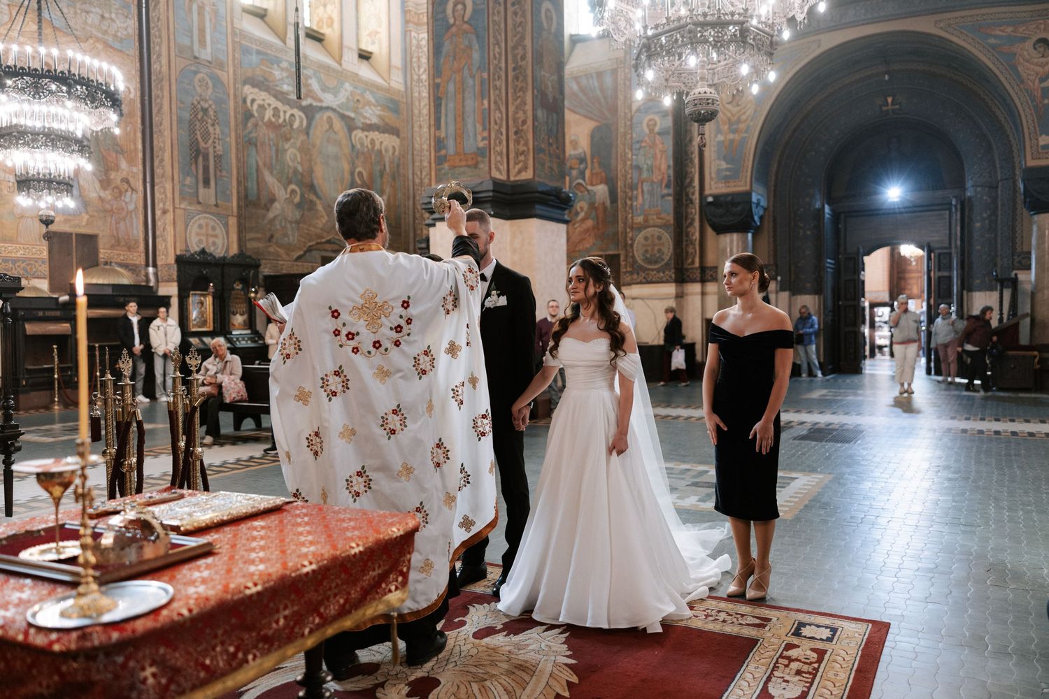 Wedding ceremony taking place in ornate orthodox church with decorated walls and chandelier.