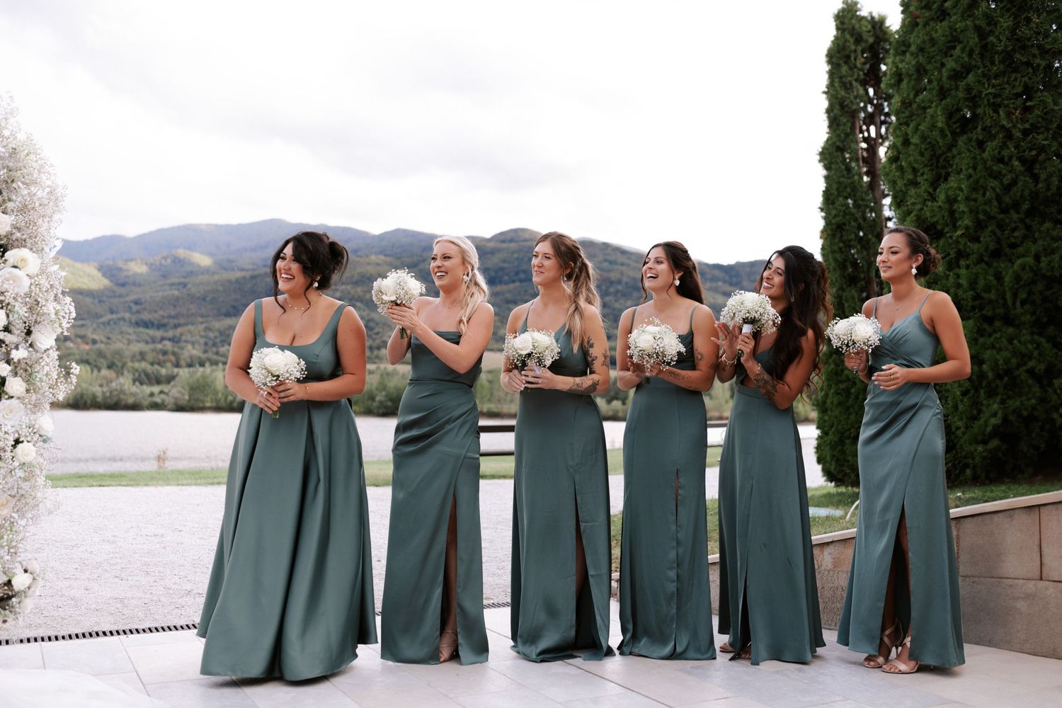 Row of bridesmaids in sage green dresses pose on mountain terrace.