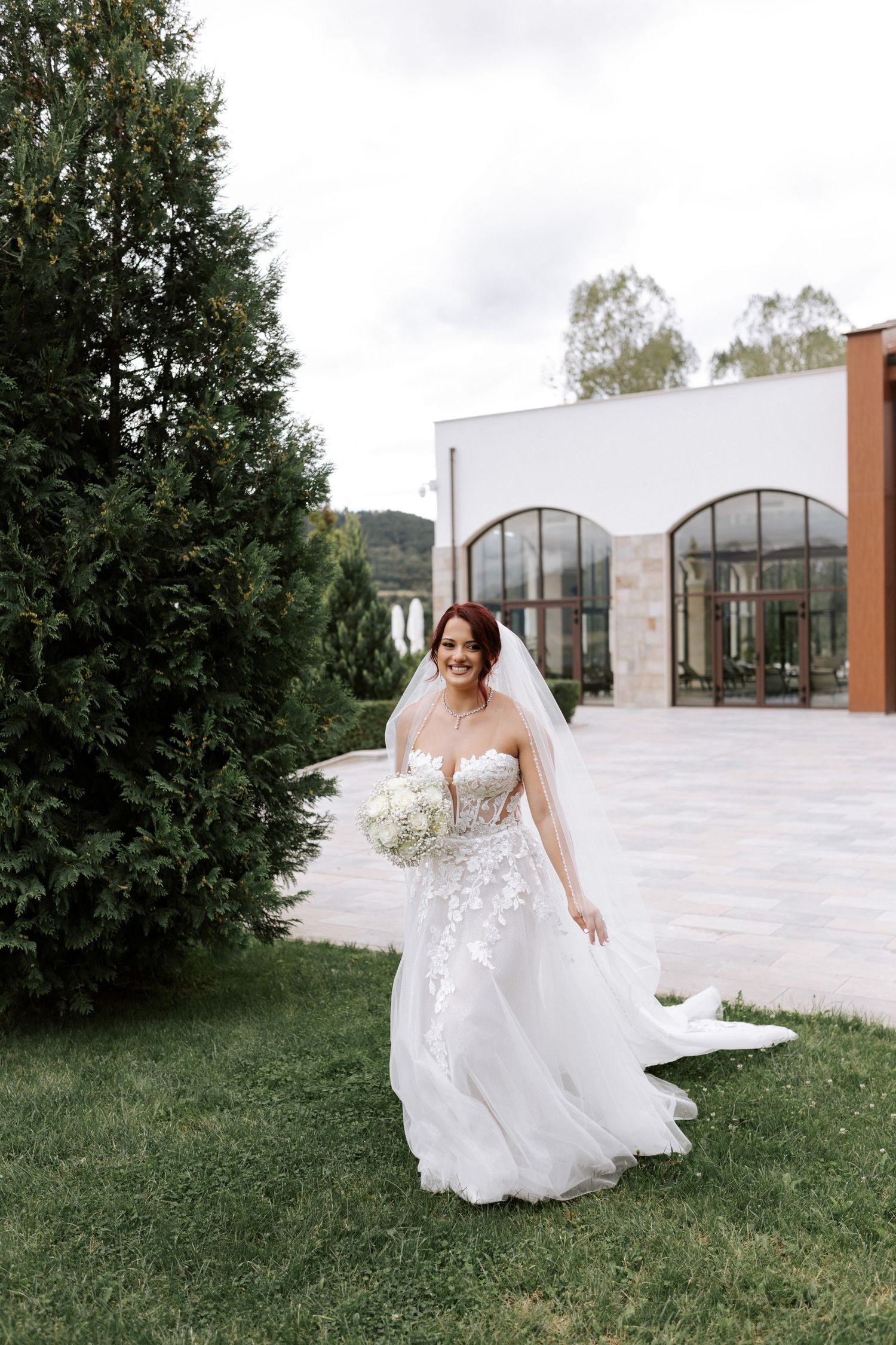 Bride in flowing white lace dress poses in front of modern venue with mountain backdrop.