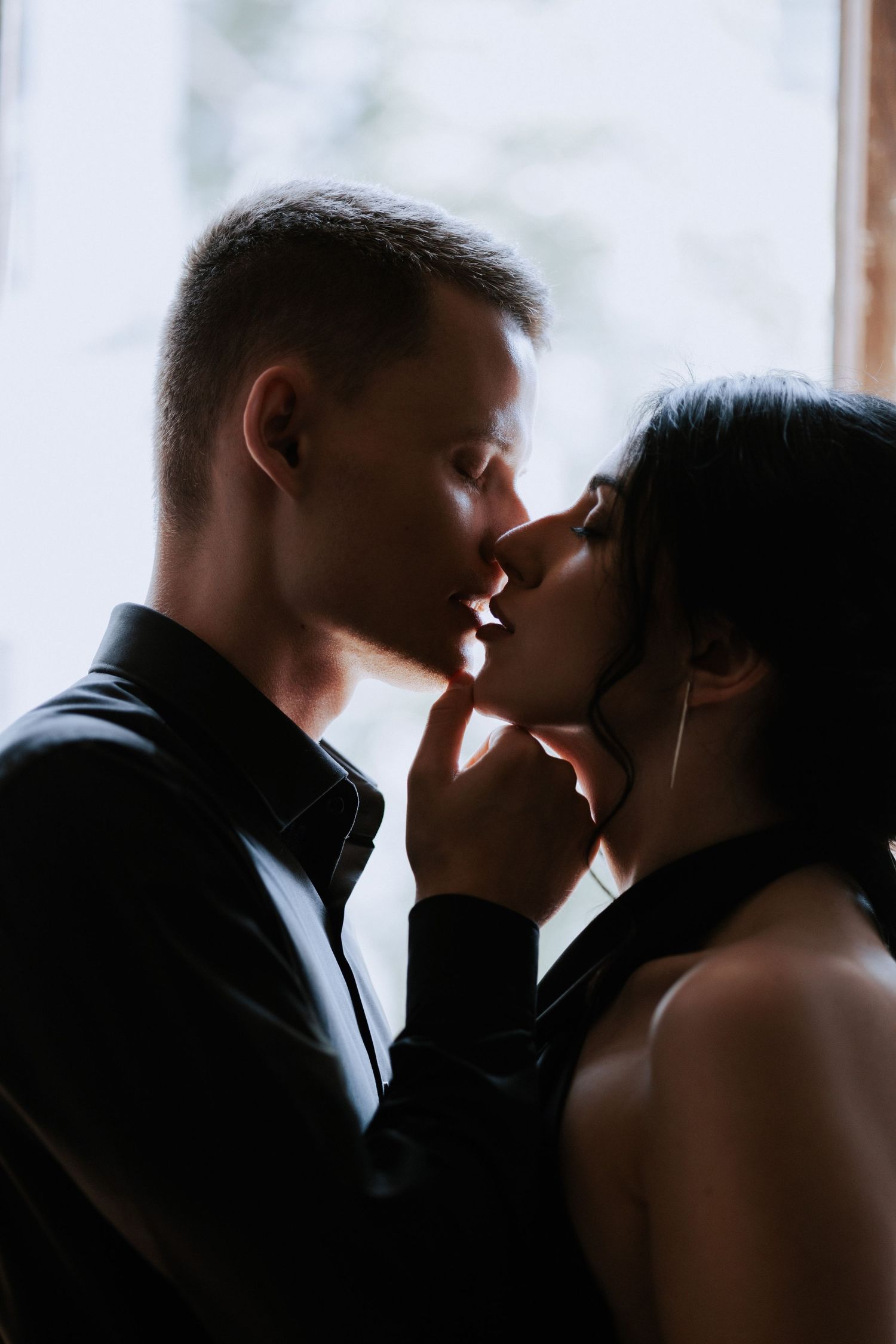Silhouetted couple share intimate moment near window in soft lighting.
