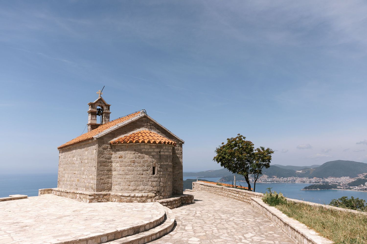 Small stone chapel with bell tower sits atop coastal cliff overlooking Mediterranean bay and mountains in distance.