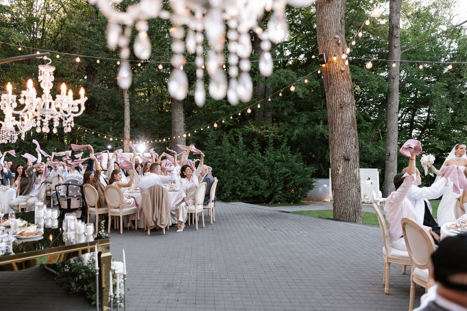 Outdoor wedding reception setup with string lights and elegant white decor under tall trees.