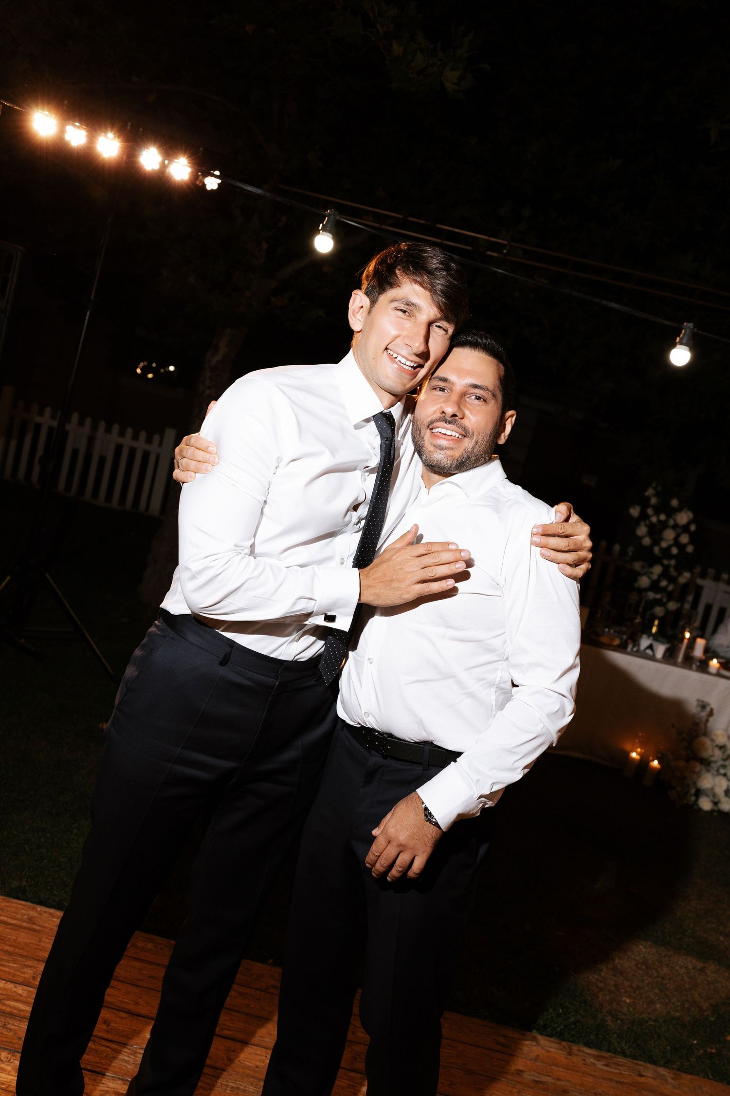 Two people in white dress shirts and black pants share playful candid moments at an evening outdoor event.