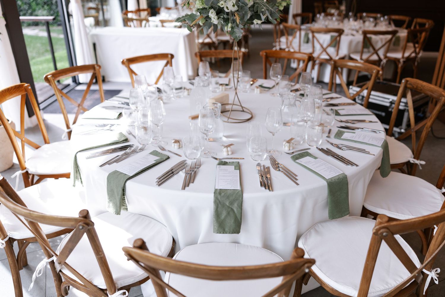Wedding reception venue with round tables set with white linens and wooden cross-back chairs in an elegant indoor space.