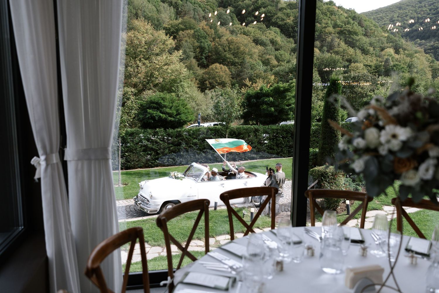 Elegant wedding reception table setup with vintage car visible through window overlooking green hillside.