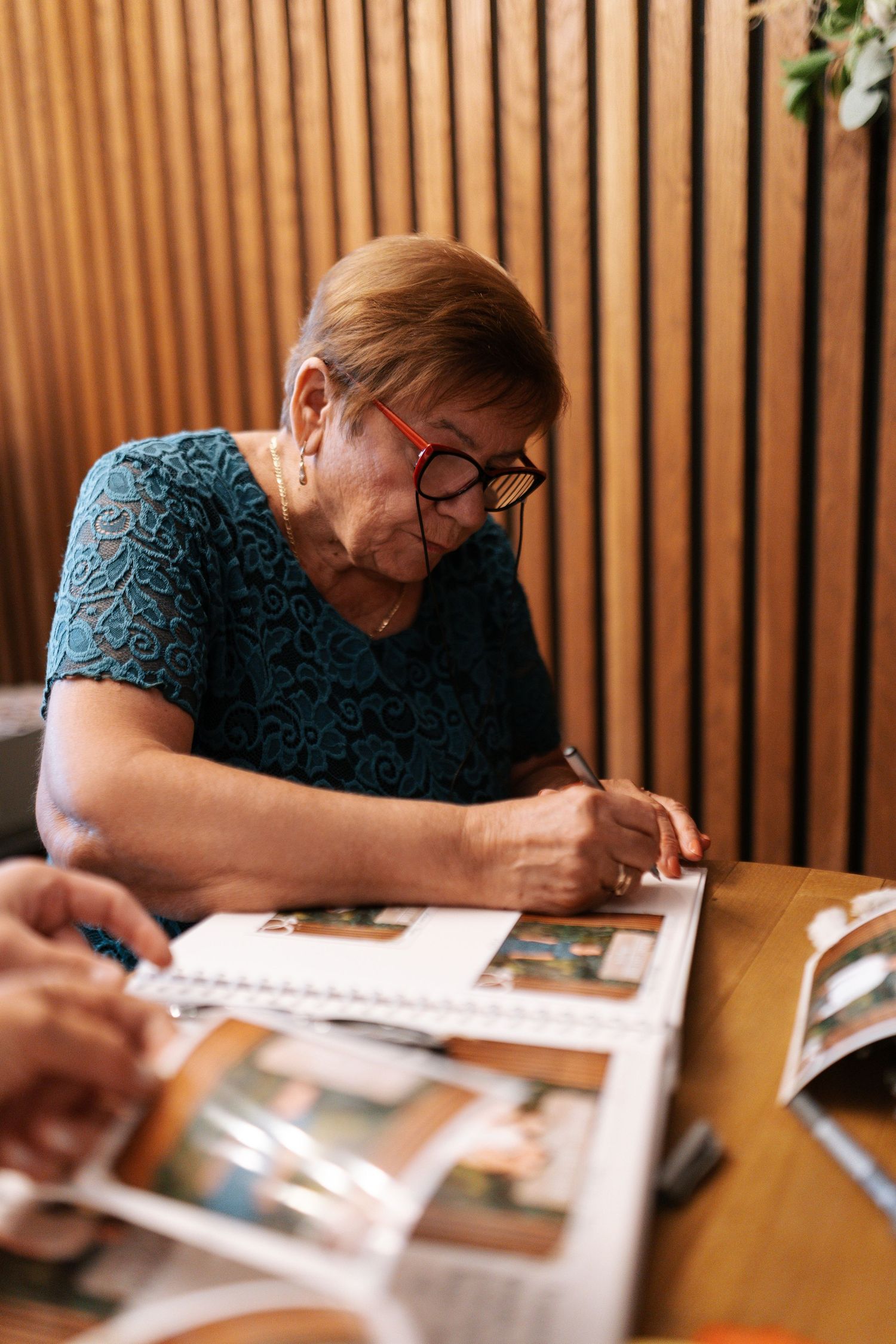 Someone wearing glasses and a blue lace top writes in a photo album at a wooden table.
