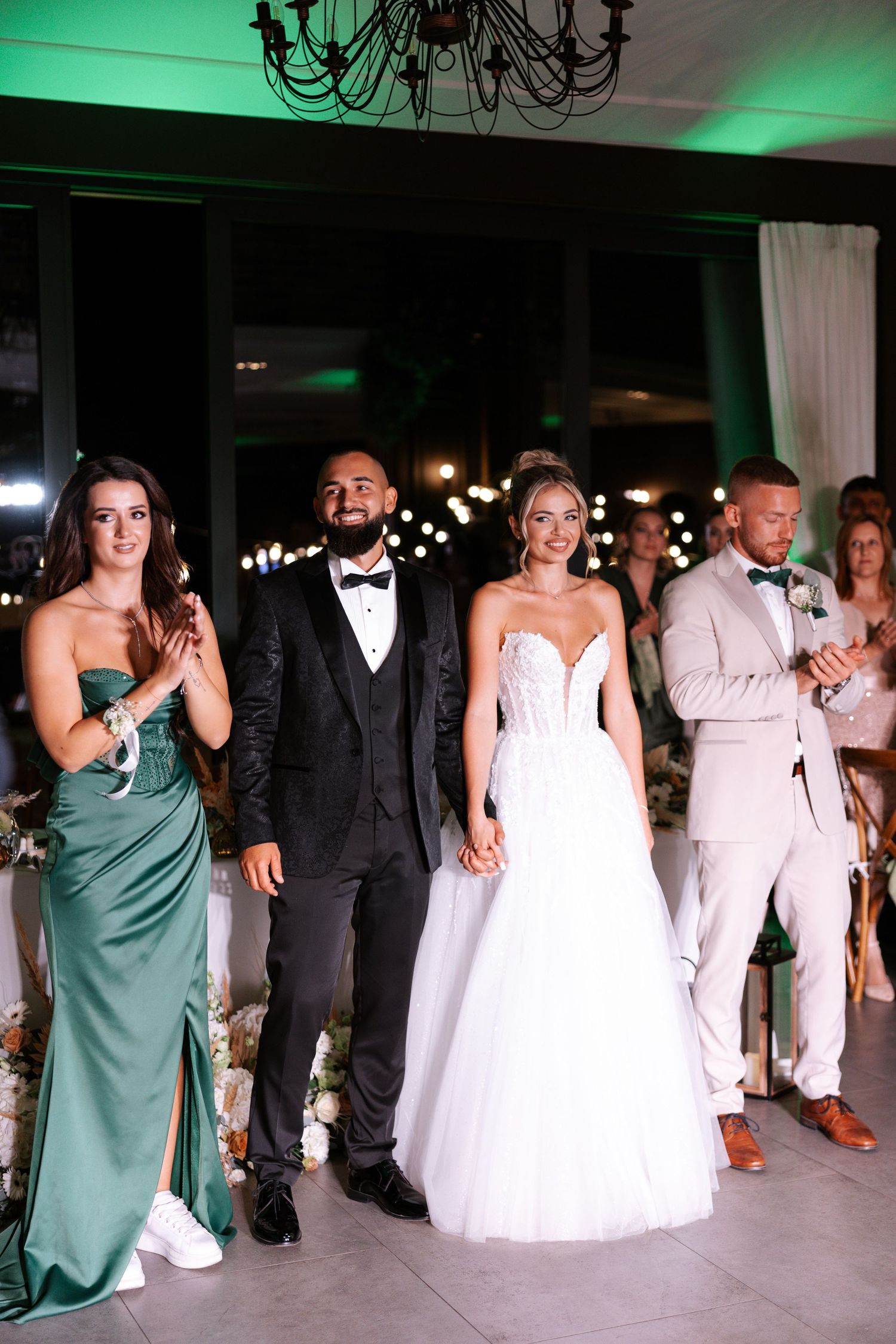 Wedding party dances in a line during reception with green uplighting and elegant chandeliers overhead.