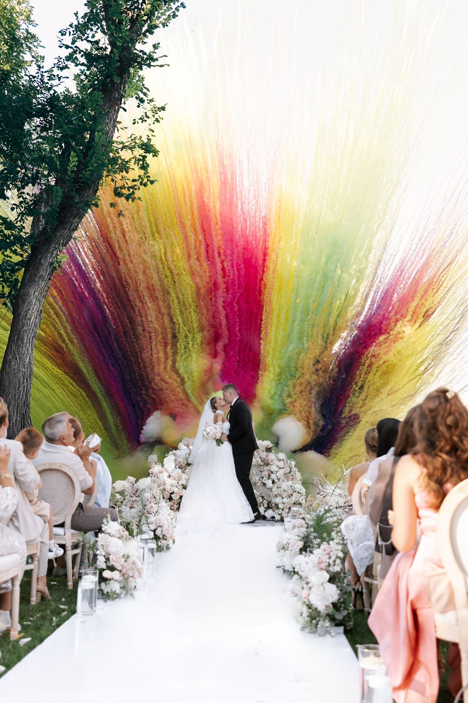 Outdoor wedding ceremony with dramatic colorful smoke bomb effect creating rainbow backdrop behind white floral aisle decor.