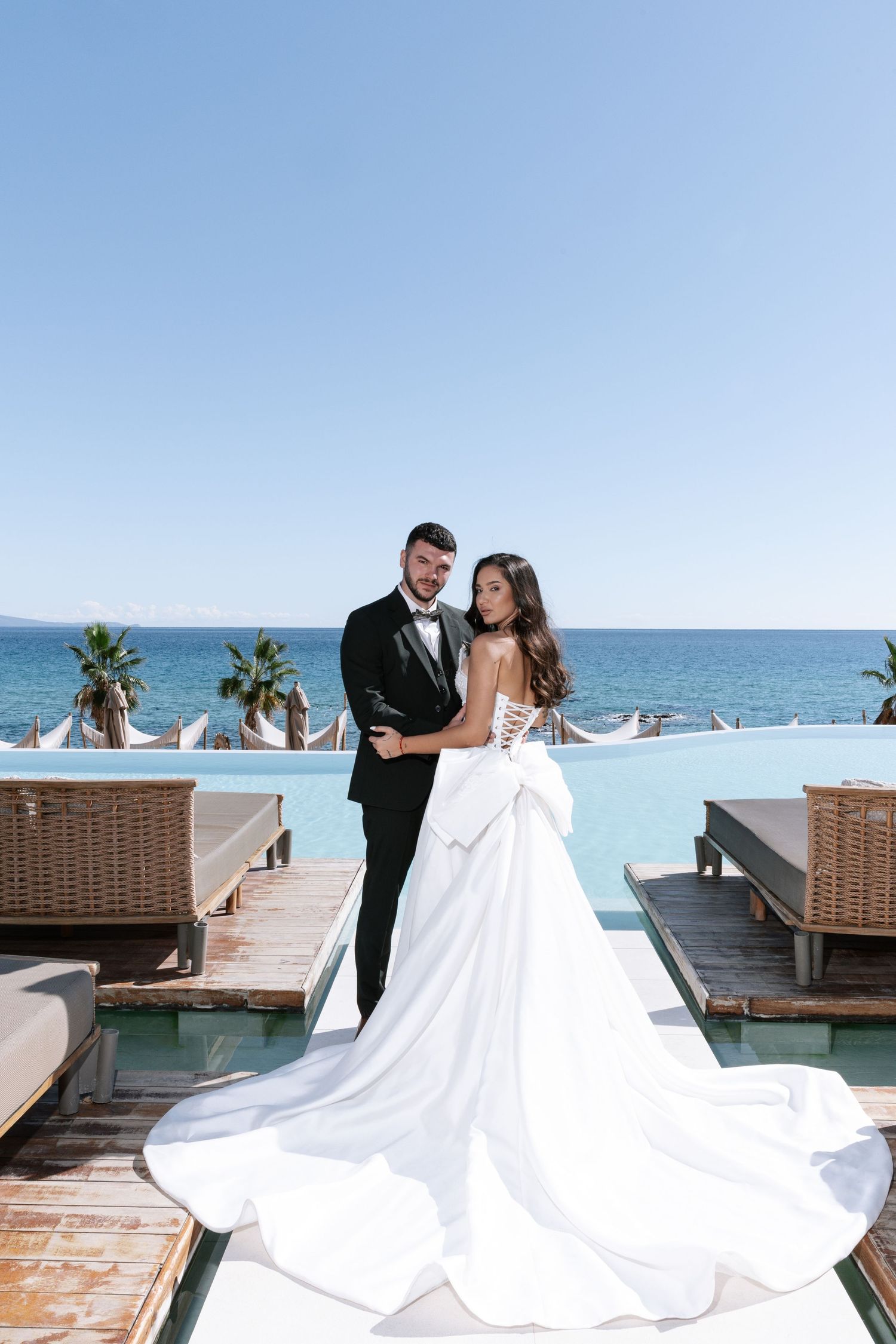 A couple shares a romantic moment on a beachfront deck with lounge chairs and palm trees in the background.