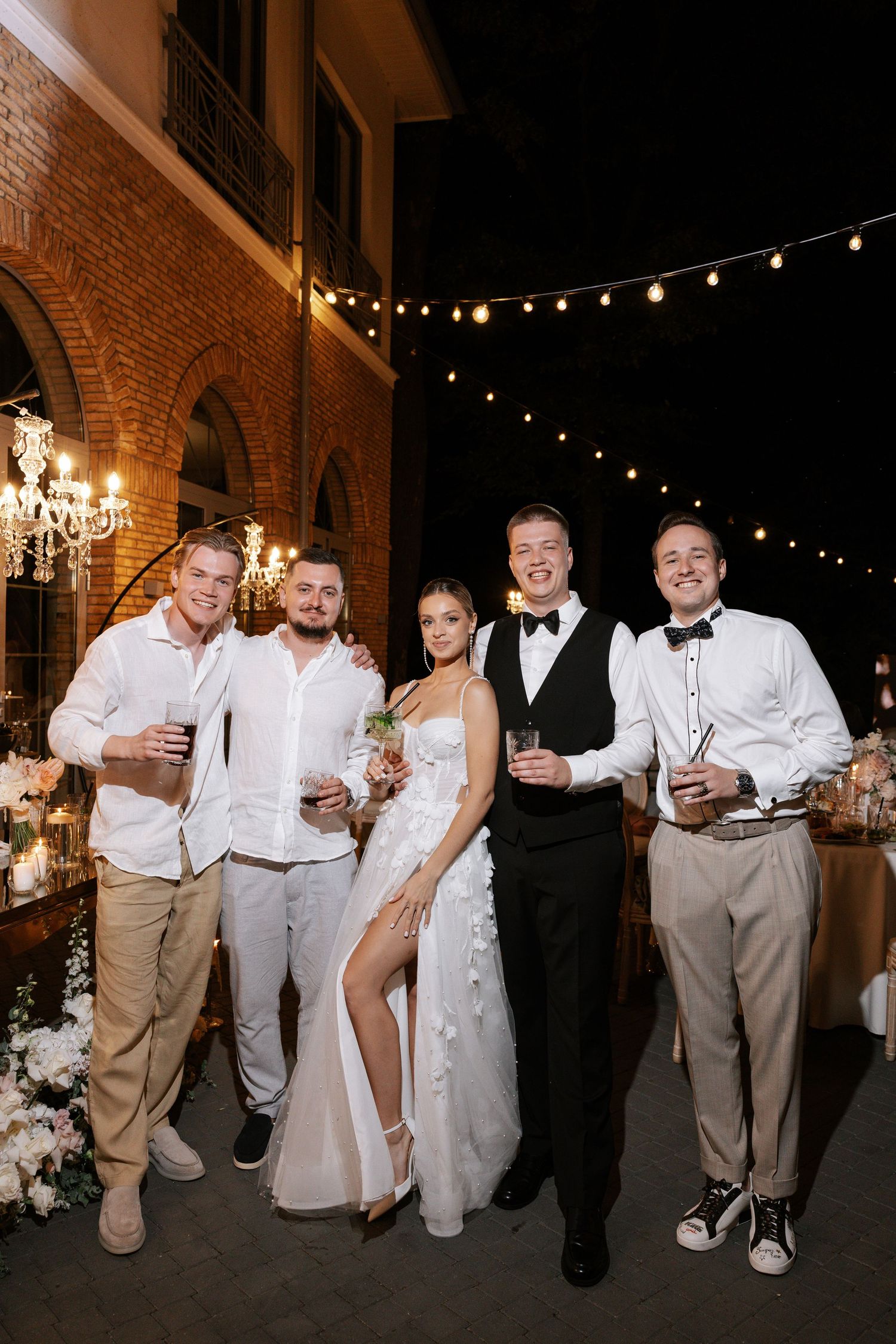 Wedding party poses together at night under string lights and brick archways.