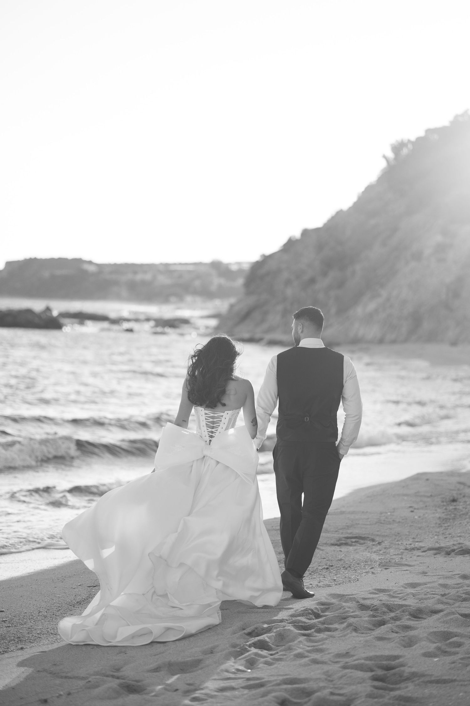 A couple walks along a misty beach at sunset with a flowing white wedding dress trailing behind them in the sand.