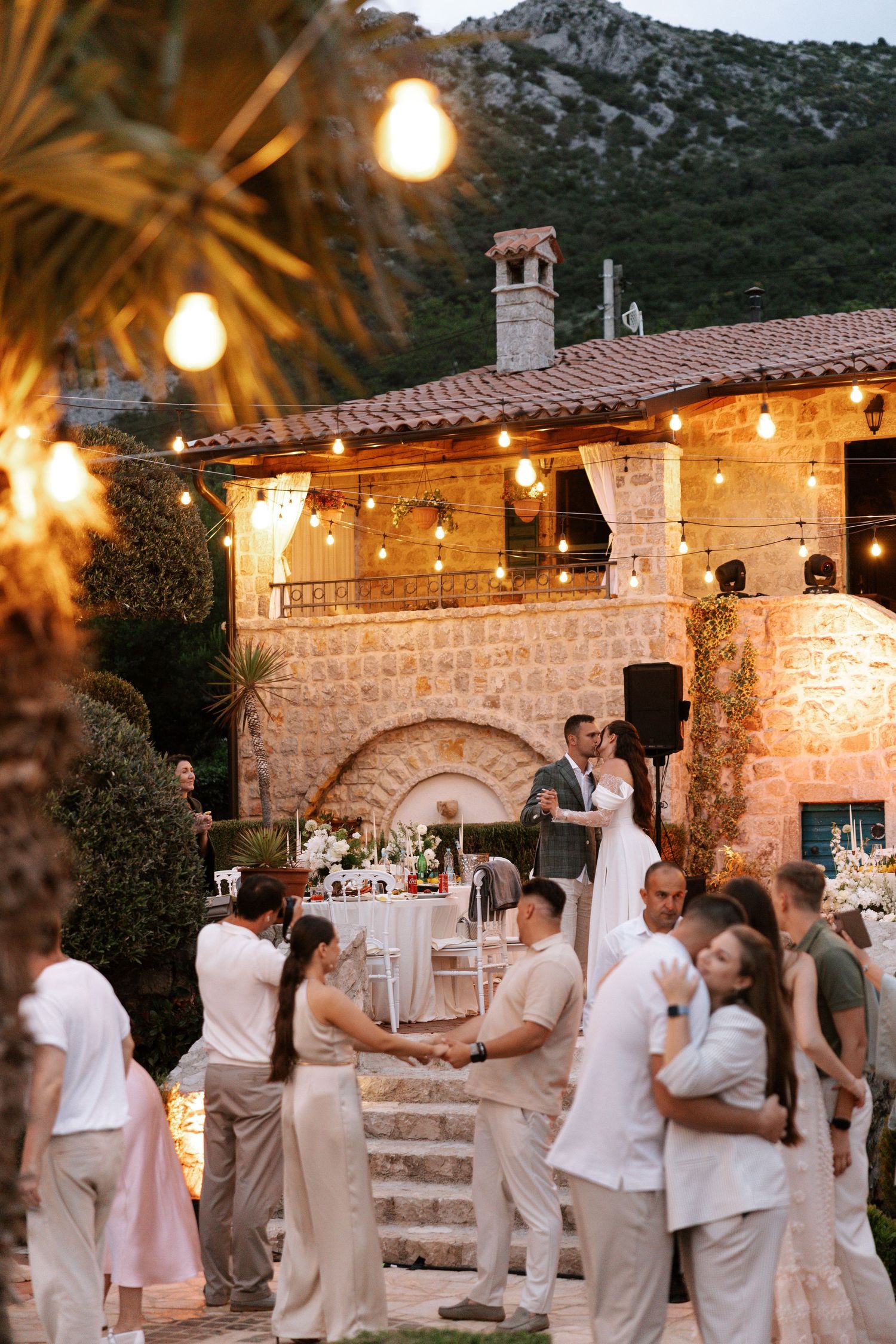 Wedding guests dance and celebrate at a rustic stone venue decorated with warm string lights at dusk.