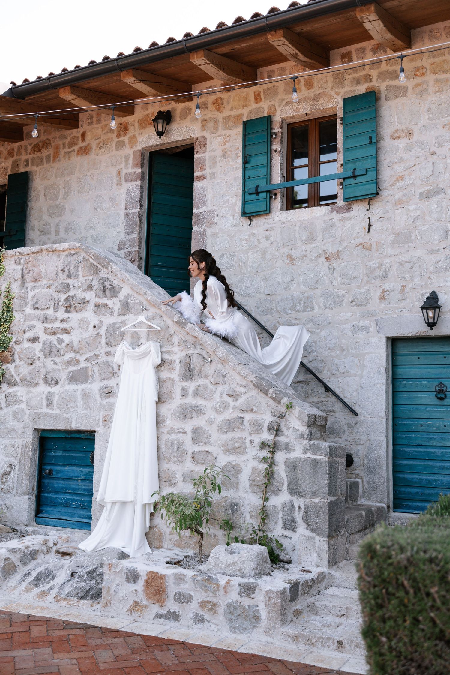 White wedding dress displayed against rustic stone wall with teal shutters and wooden beams in Mediterranean setting.