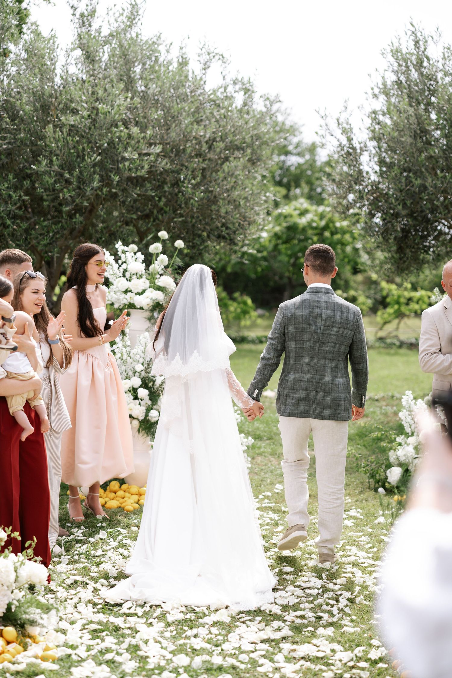 Wedding guests mingle in a sunny garden setting with scattered white flower petals and blooming trees.