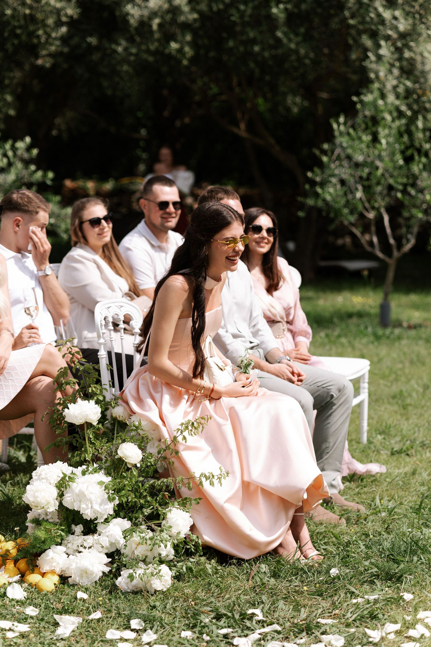 Wedding guests in light colored attire sit on white chairs outdoors during a sunny garden ceremony.