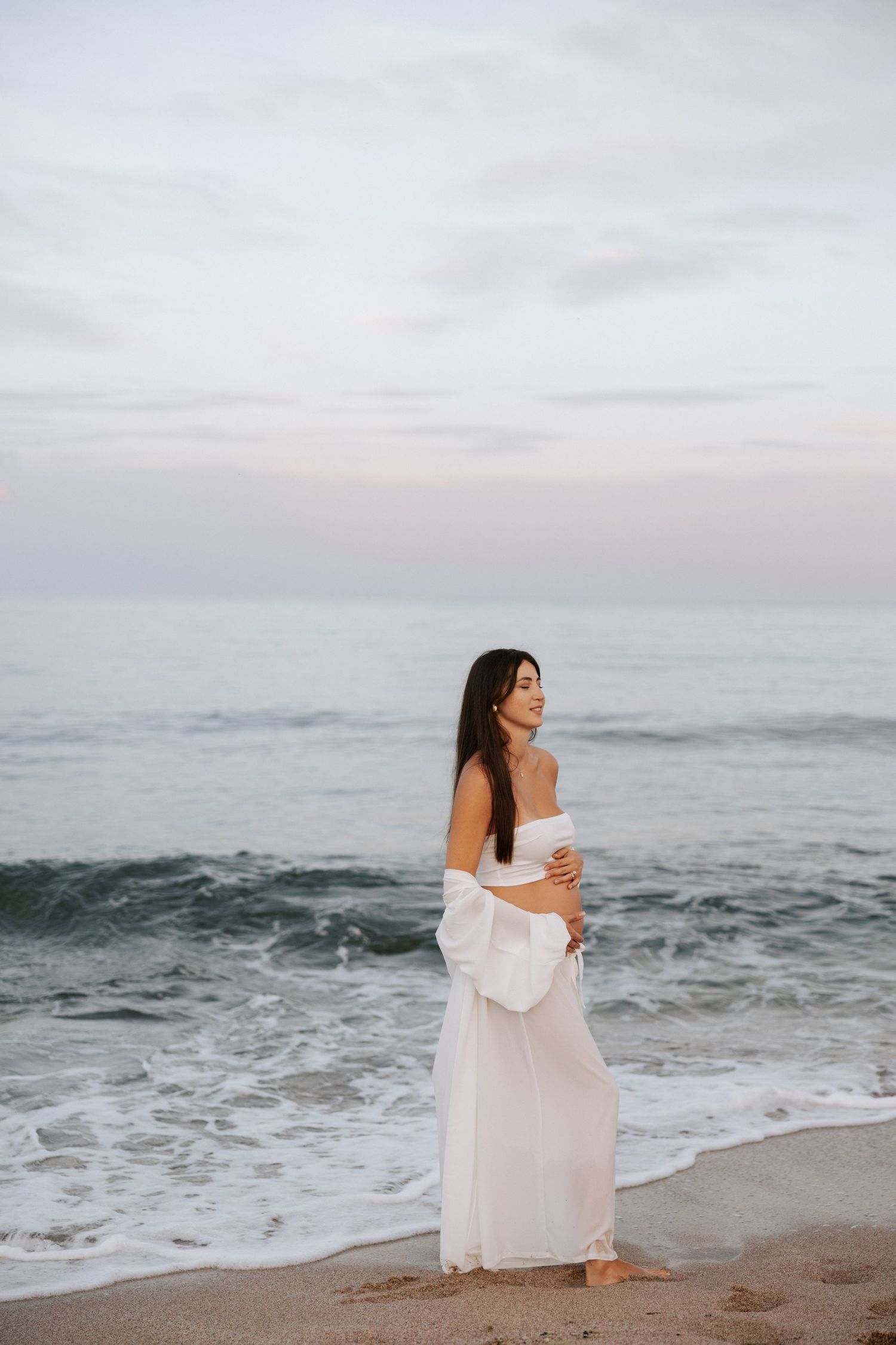 A serene maternity photo of an expectant mother in white dress standing by ocean waves.