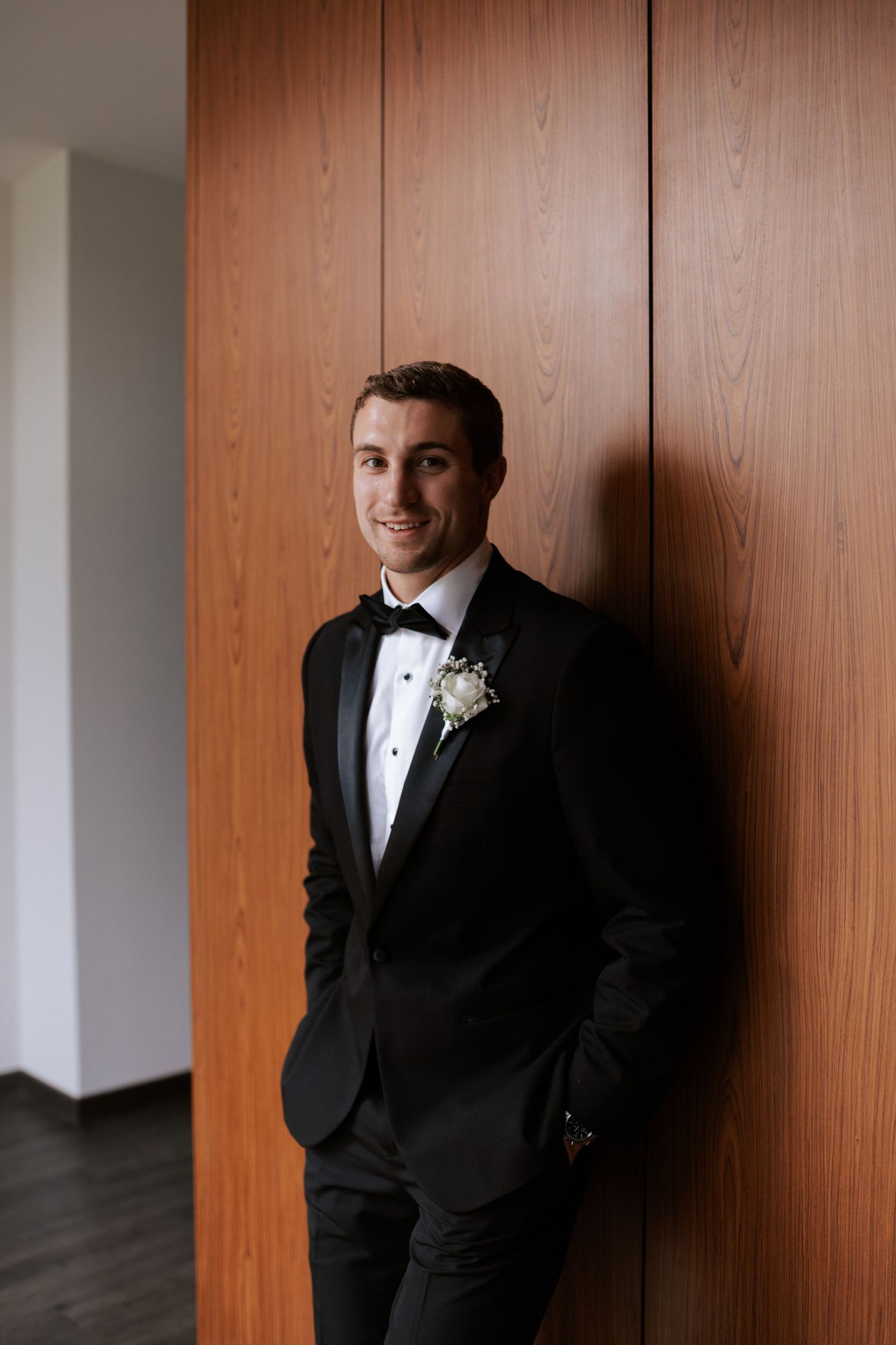 A groom in classic black tuxedo poses against wooden paneled wall.