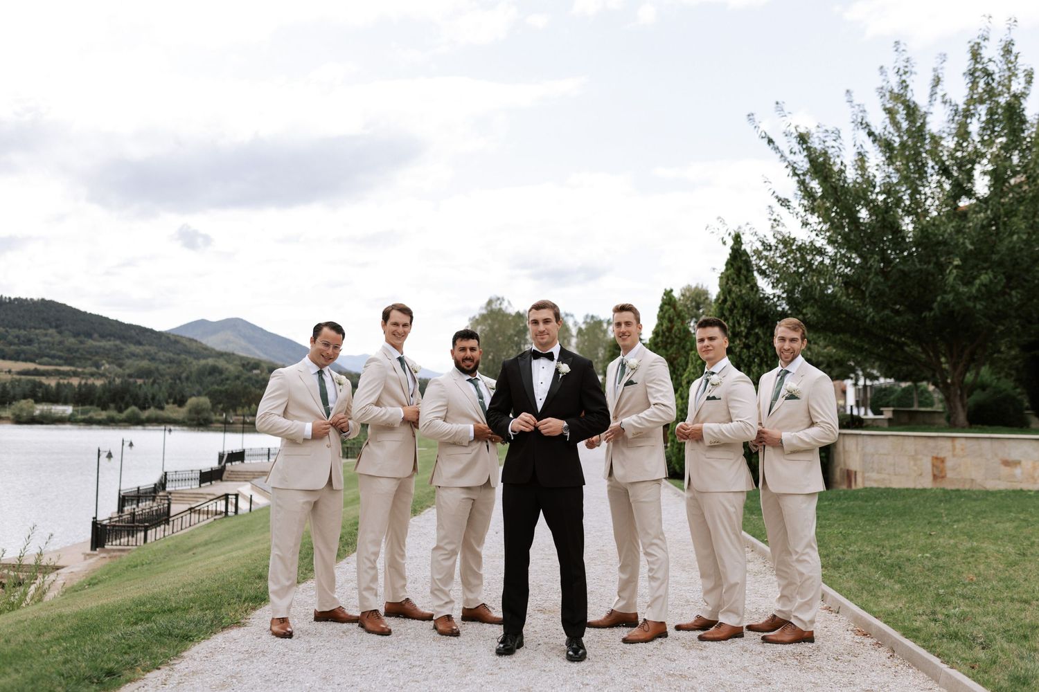 Wedding party in cream suits with one in black pose against mountain lake backdrop.
