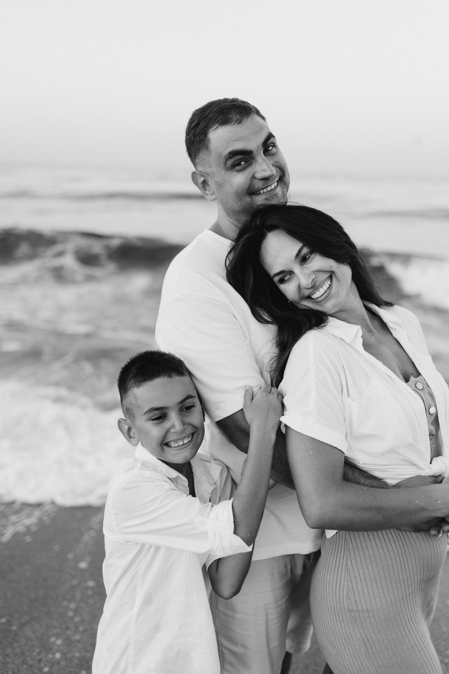A black and white beach portrait of a family embracing and smiling together.