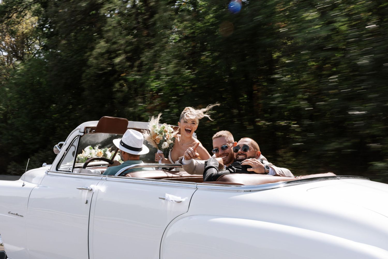 A white vintage convertible car drives through lush greenery with passengers enjoying a sunny day ride.